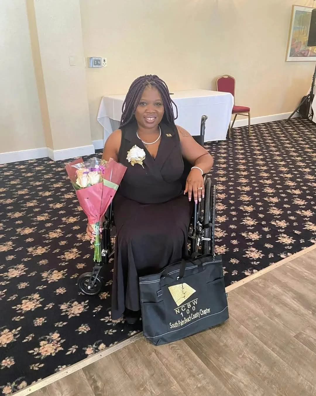 Woman in a wheelchair holding a bouquet of flowers, smiling, in a room with patterned carpet and chairs.