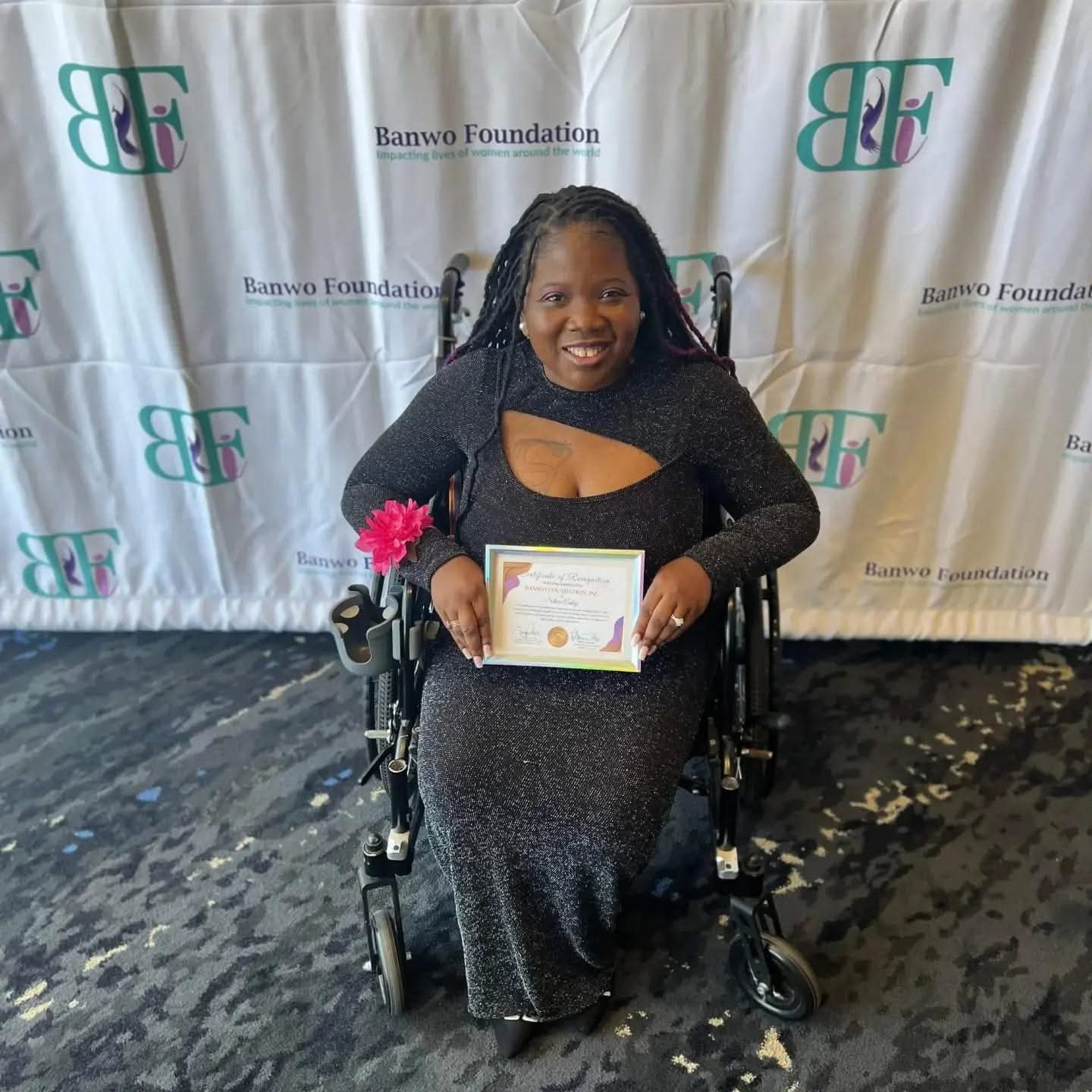 Woman in a wheelchair holding a certificate, smiling, in front of a backdrop with logos and text.