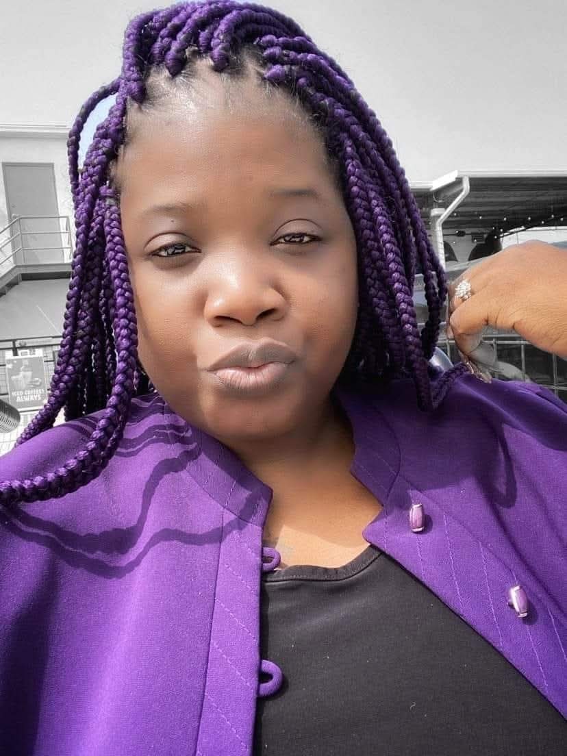 Young woman with purple braided hair taking a selfie outdoors, wearing a purple jacket and black top.