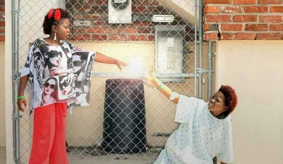 Two children interact through a chain-link fence, one standing and pointing, the other sitting with a hand extended holding a light.