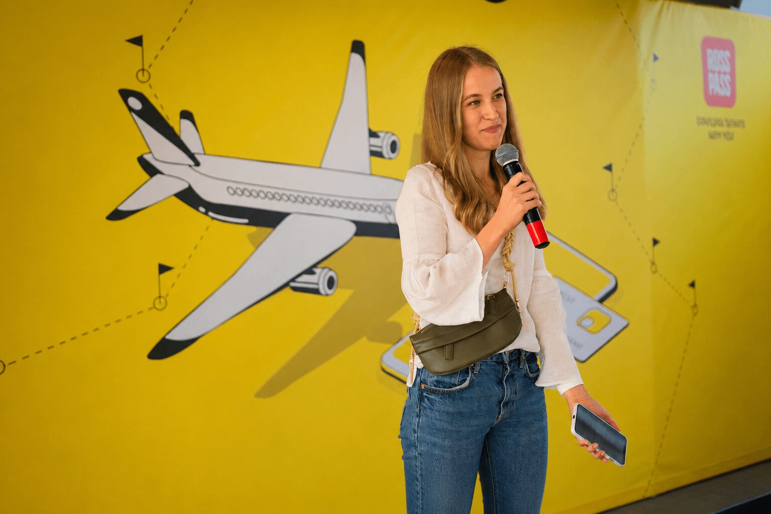 Young woman holding a microphone and a booklet, standing in front of a yellow wall with an airplane illustration.