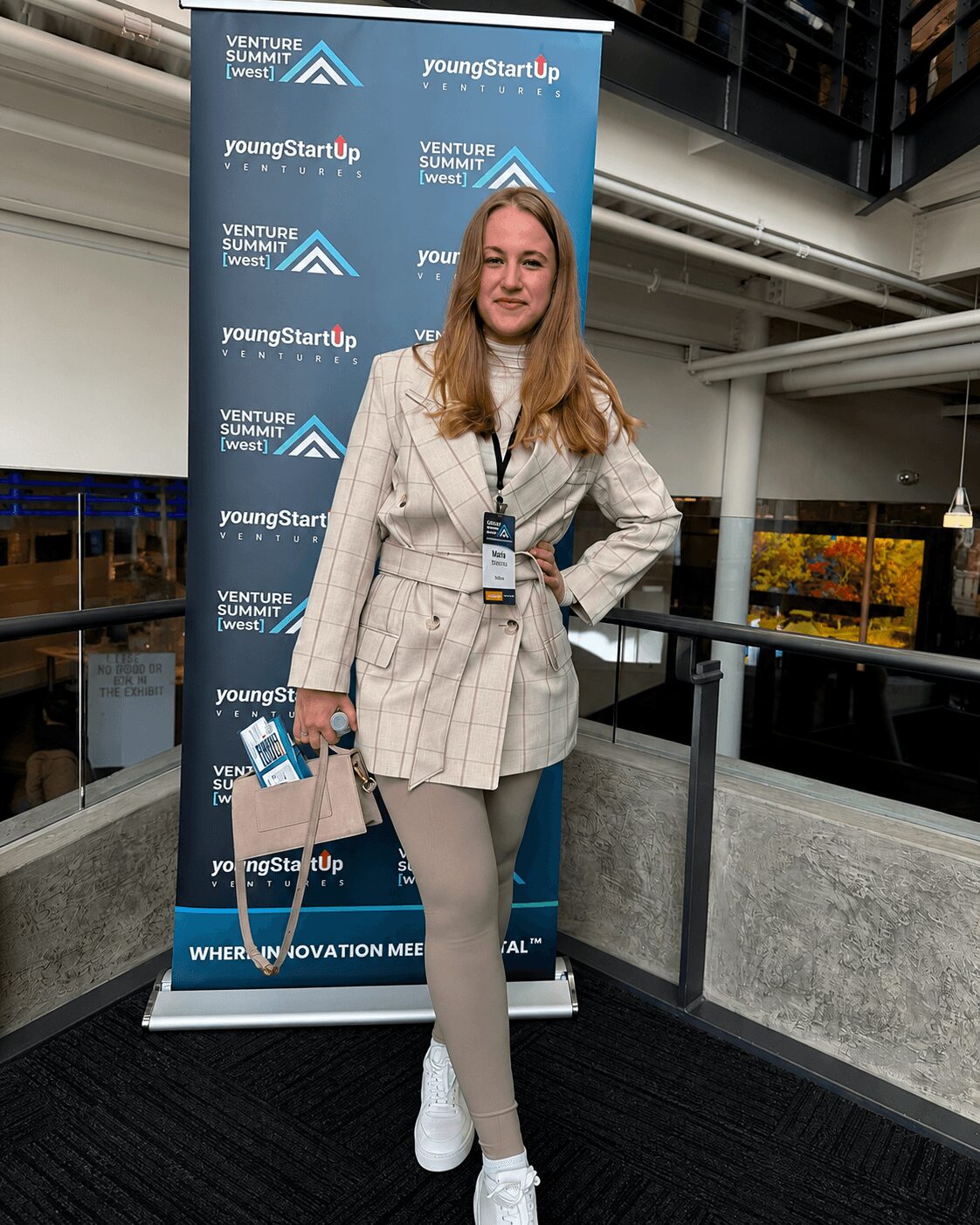 Woman standing in front of a blue event backdrop, smiling, wearing a beige blazer, white sneakers, and holding a bag.