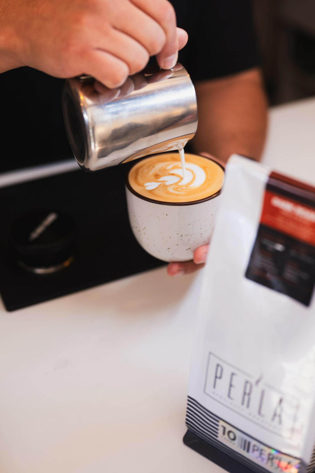 Barista pours steamed milk into a cup of coffee with latte art, on a white surface, with coffee bag nearby.
