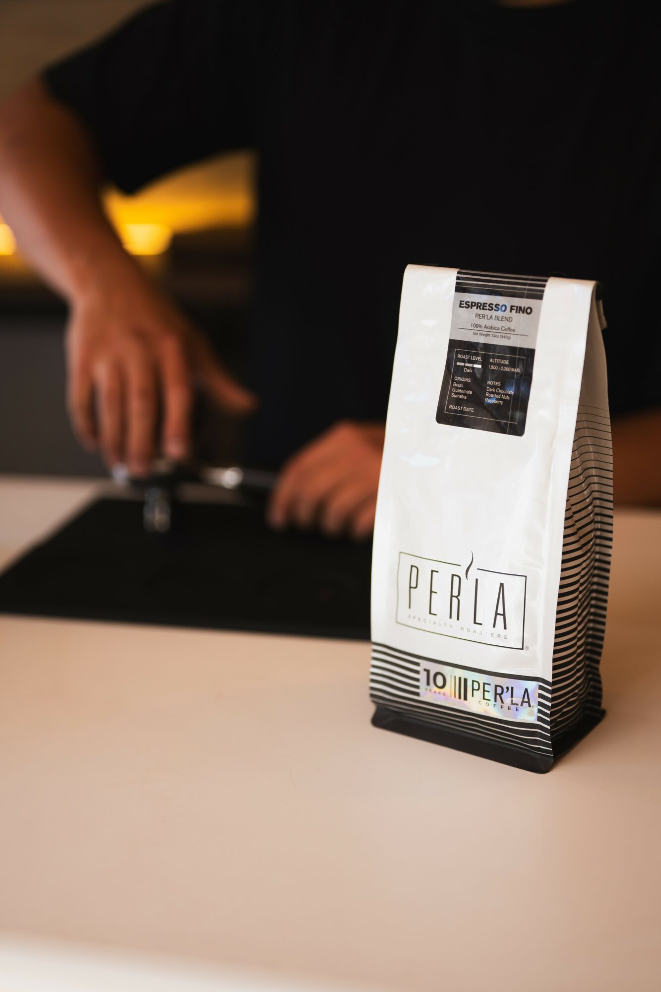 White paper bag with black text reading 'PERLA' on a white surface, with a person in black shirt in the background.