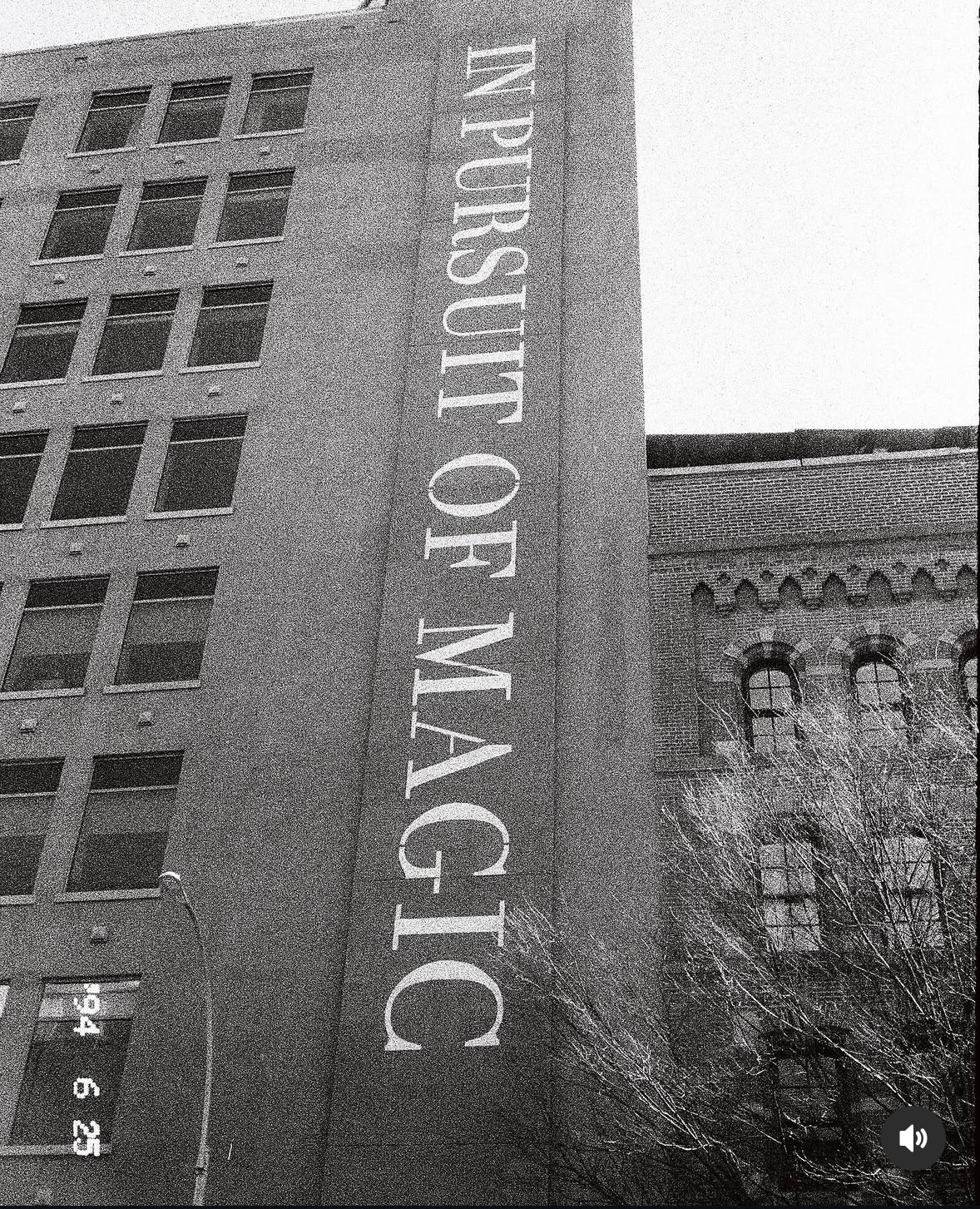 Building with vertical sign reading 'IN PURSUIT OF MAGIC' on the side, black and white photo, multiple windows, trees in foreground.