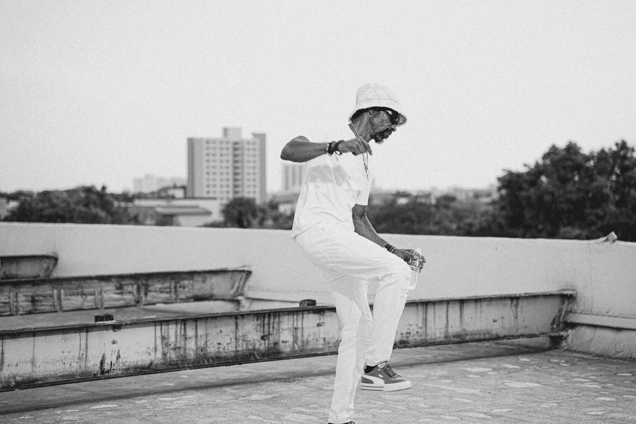 Person in white outfit and hat sitting on a rooftop ledge, city buildings in background, black and white photo.