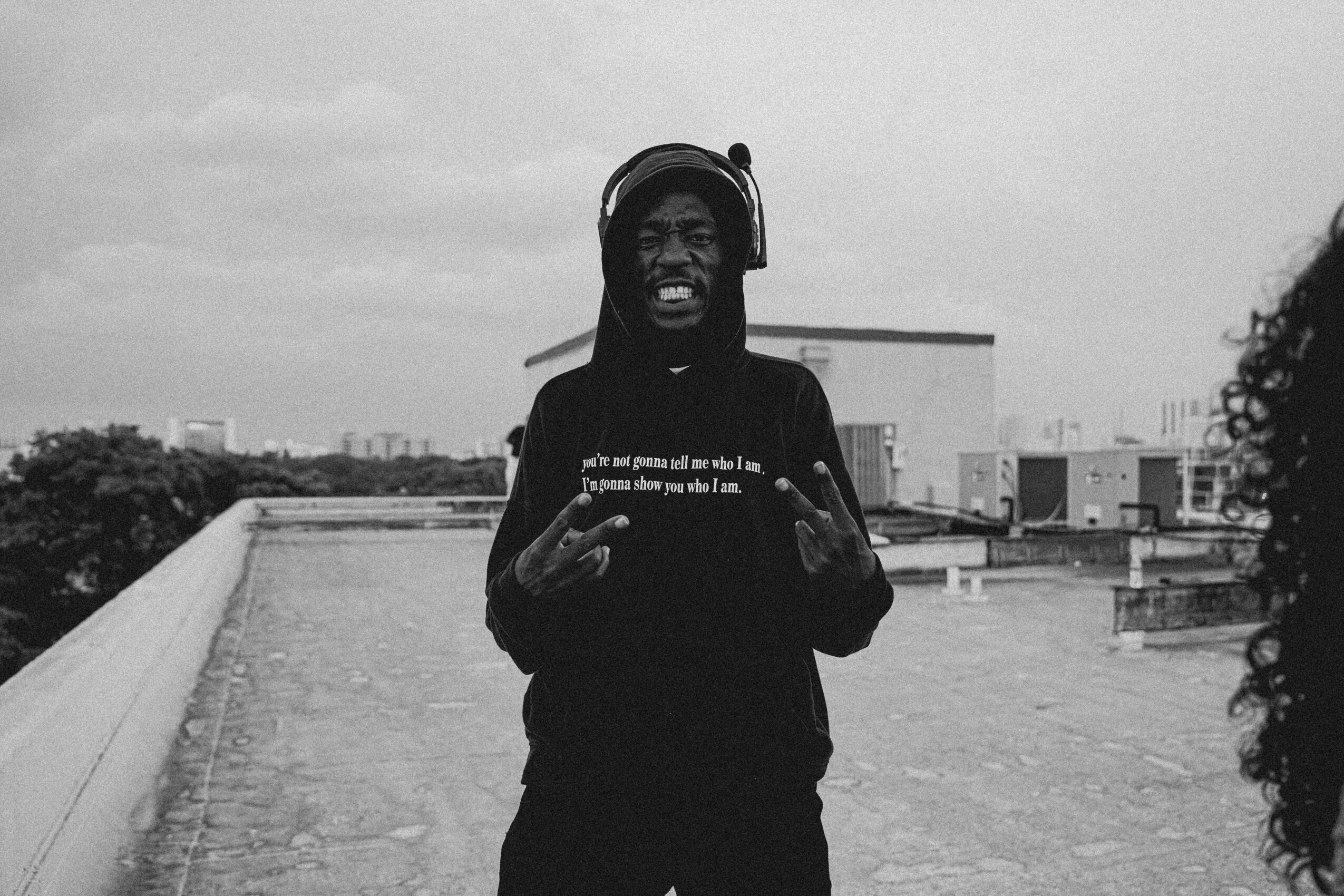Person wearing headphones, smiling, standing outdoors on a rooftop with cityscape in background.