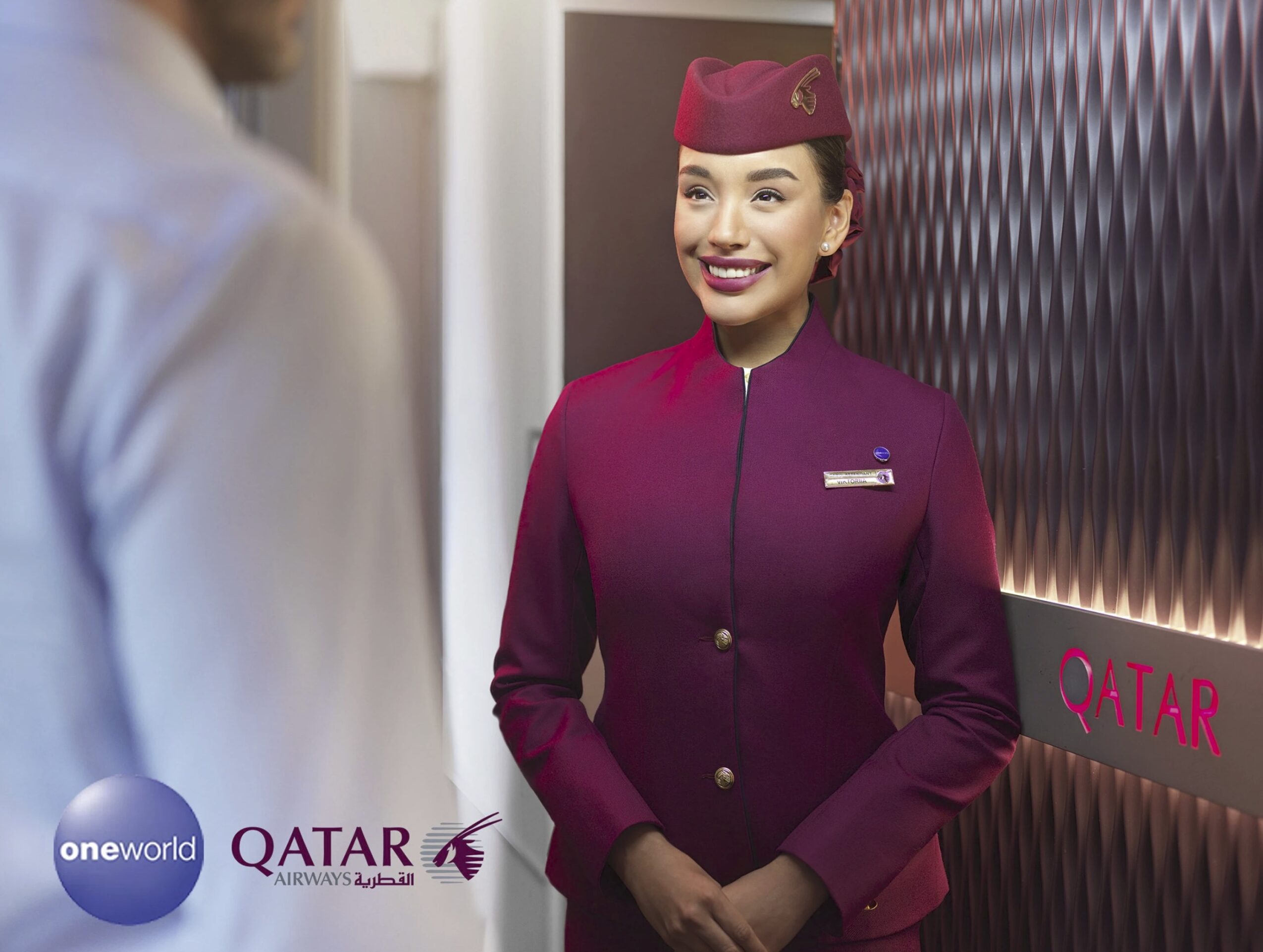 Female airline staff in uniform smiling at a passenger, standing near a Qatar Airways sign, indoors.