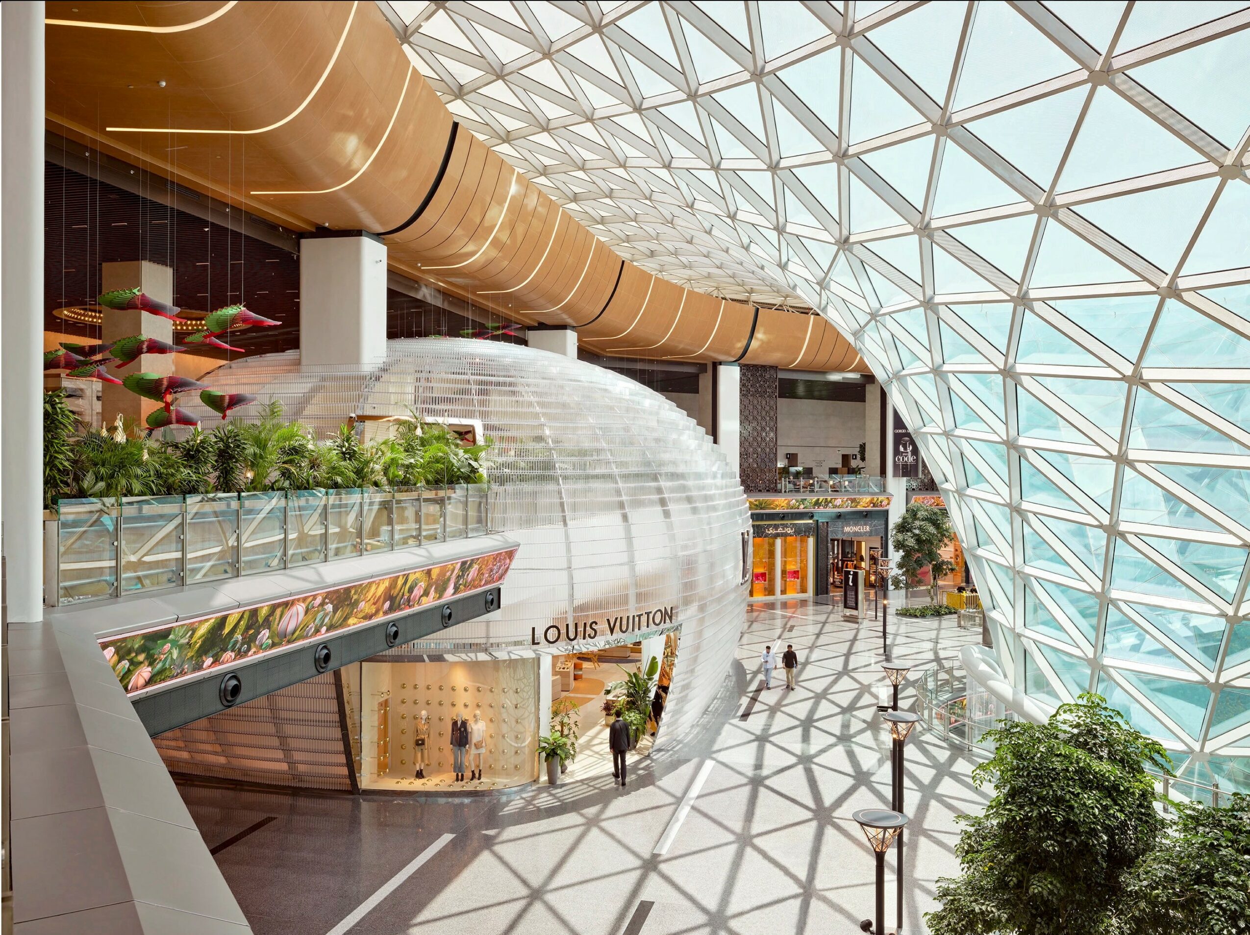 Interior of a modern shopping mall with glass ceiling, curved architecture, and storefronts, including Louis Vuitton, with plants and lighting.