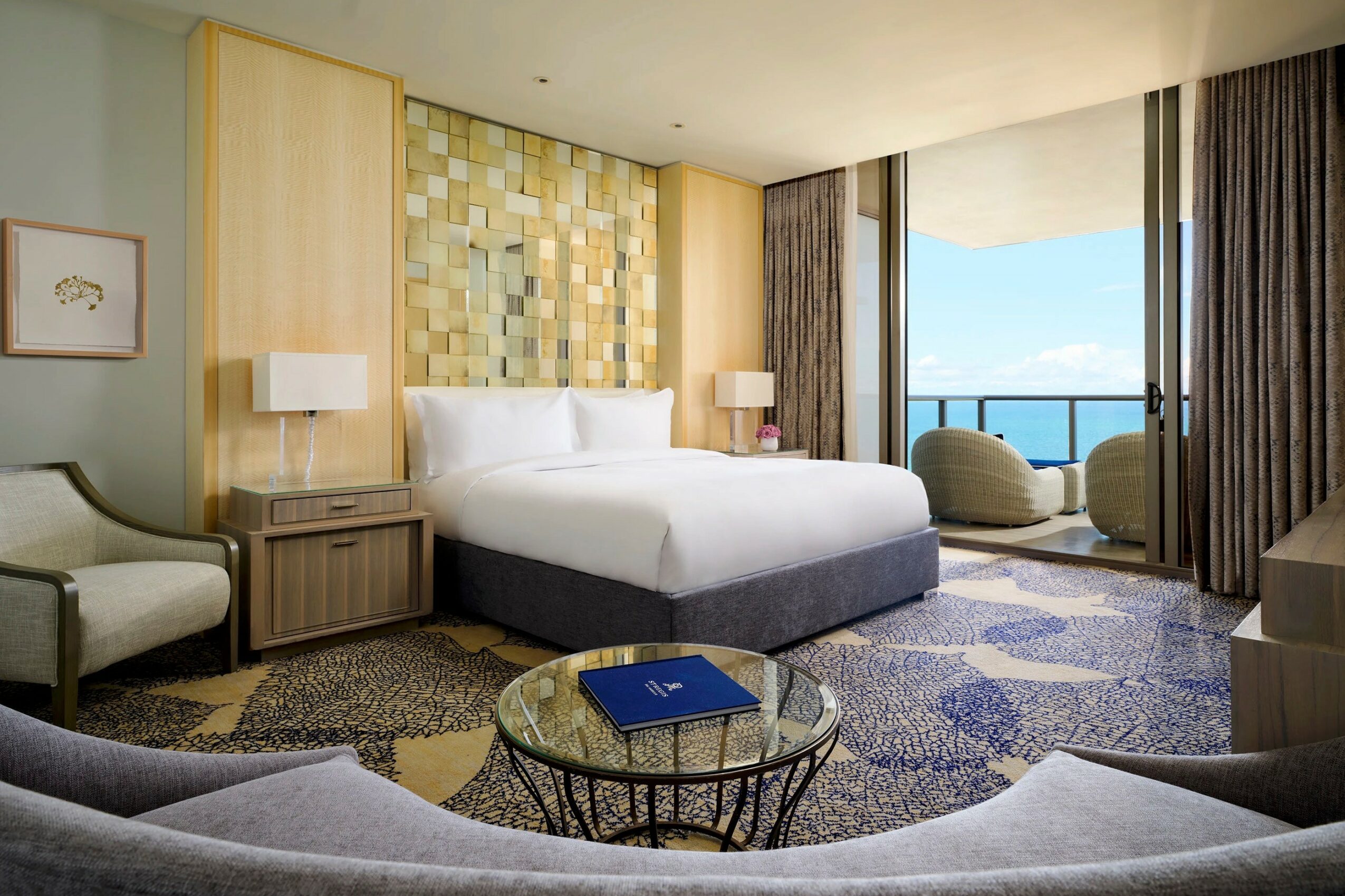 Hotel room with a bed, two nightstands with lamps, a sitting chair, and a balcony with ocean view.