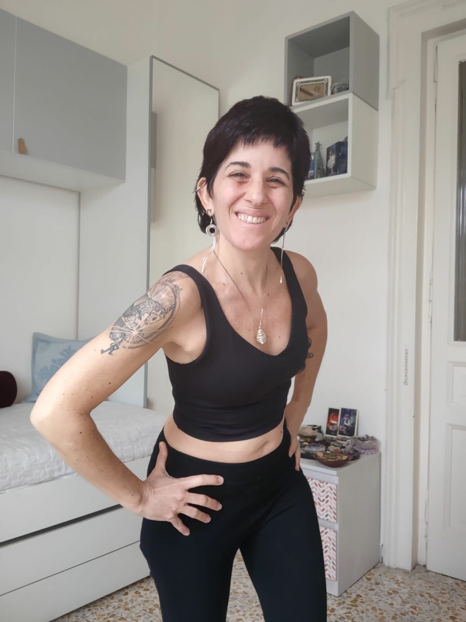 Woman smiling, standing in a room with white walls and furniture, wearing a black crop top and black pants, with tattoos on her arm.