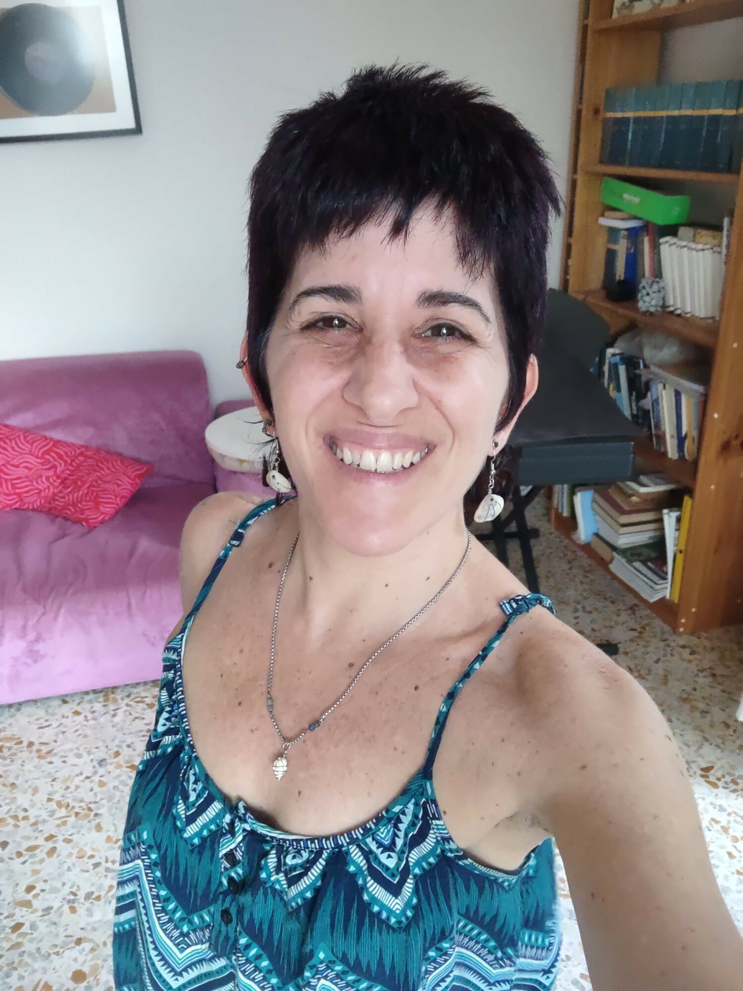 Woman with short dark hair smiling, wearing a blue patterned top, sitting on a pink couch in a room with books and a framed picture.