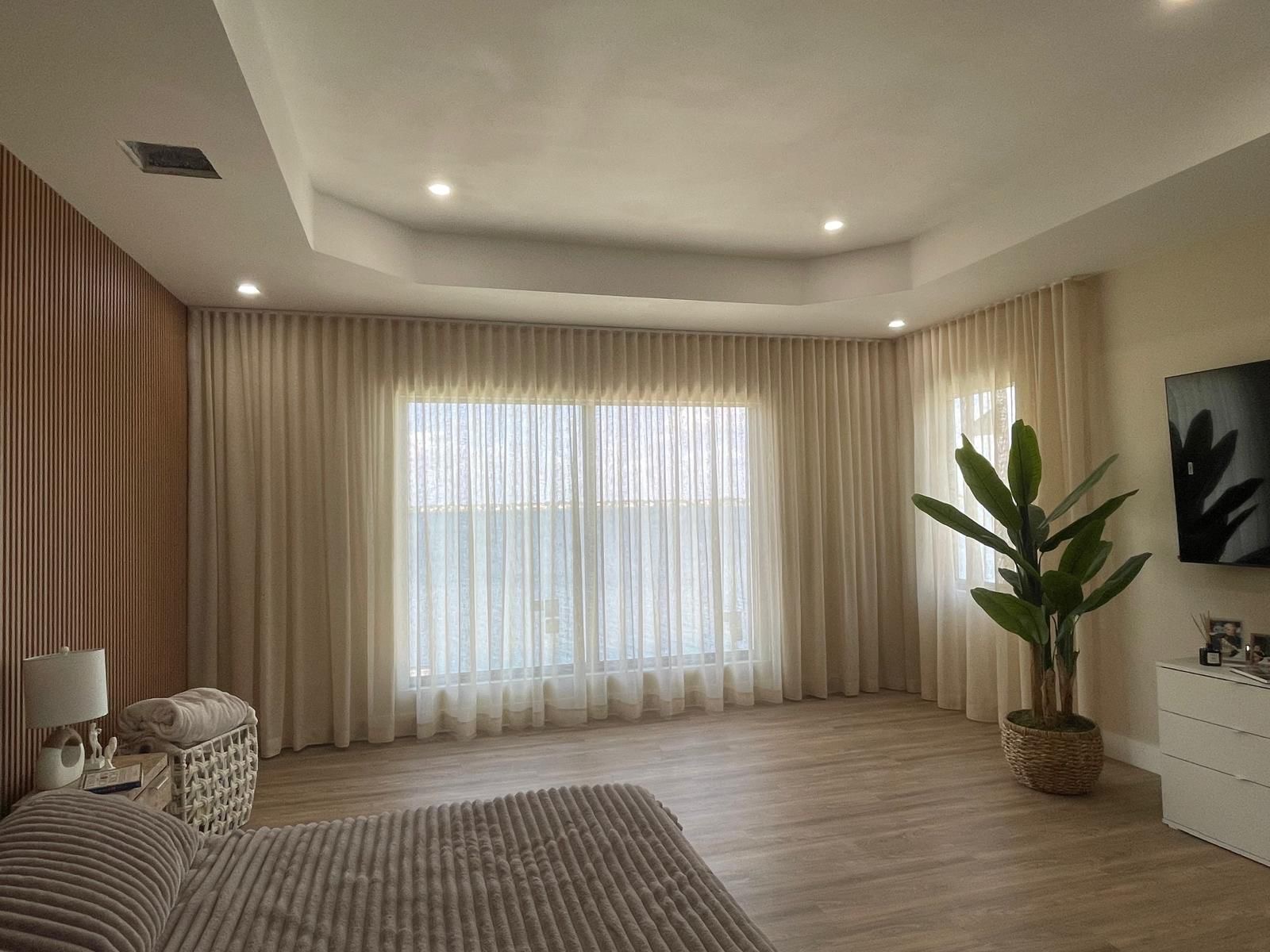 Living room with large window, beige curtains, potted plant, and modern decor, illuminated by ceiling lights.