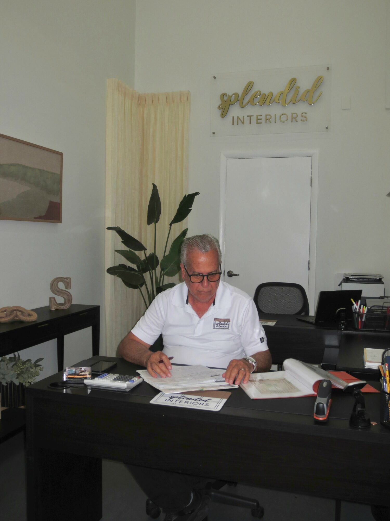 Person with glasses sitting at desk, writing on paper, in an office with a sign reading 'Glamour Interiors' on the wall.