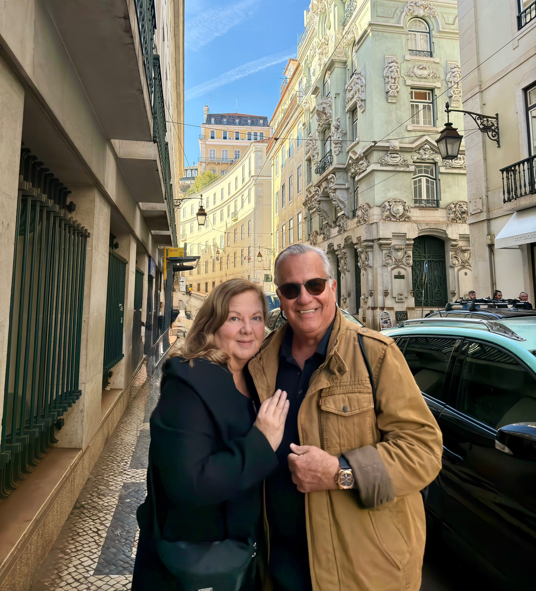 Two people smiling on a city street with historic buildings and a black car in the background.