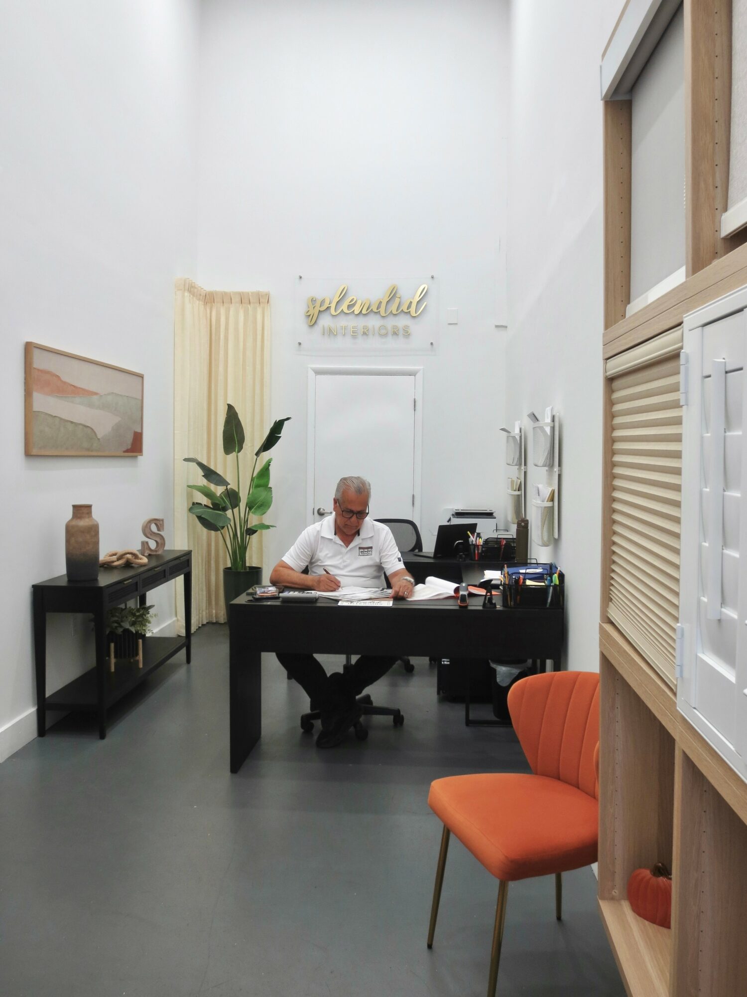Office with a person sitting at a desk, a plant, framed picture, and orange chair, with white walls and high ceiling.