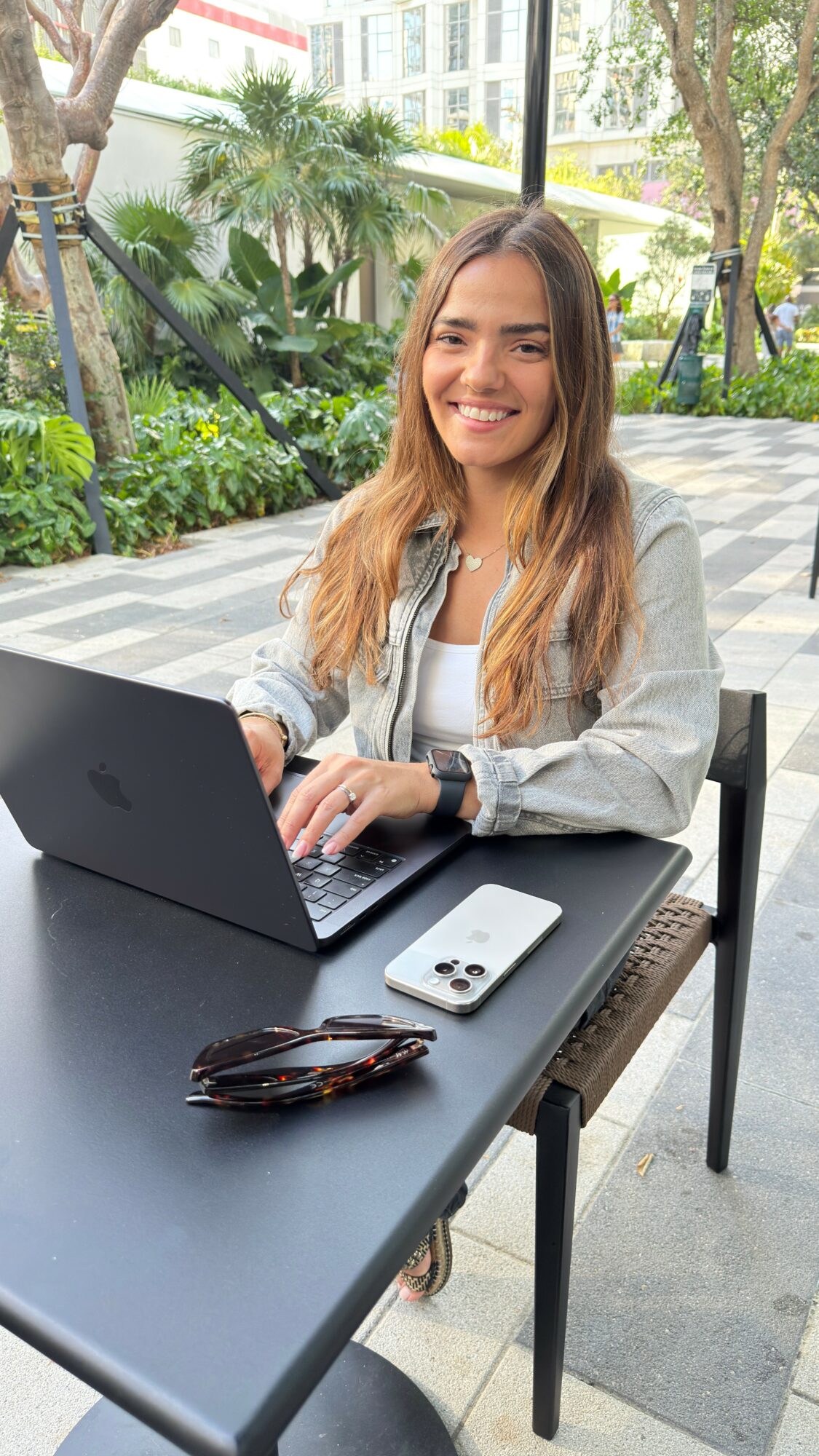 Young woman with long hair smiling at laptop outdoors, sitting at a black table with sunglasses and phone, greenery in background.