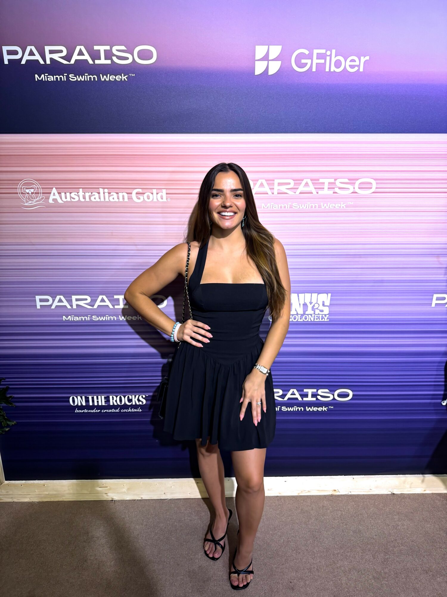 Smiling woman in a black dress standing in front of a purple backdrop with logos and text.
