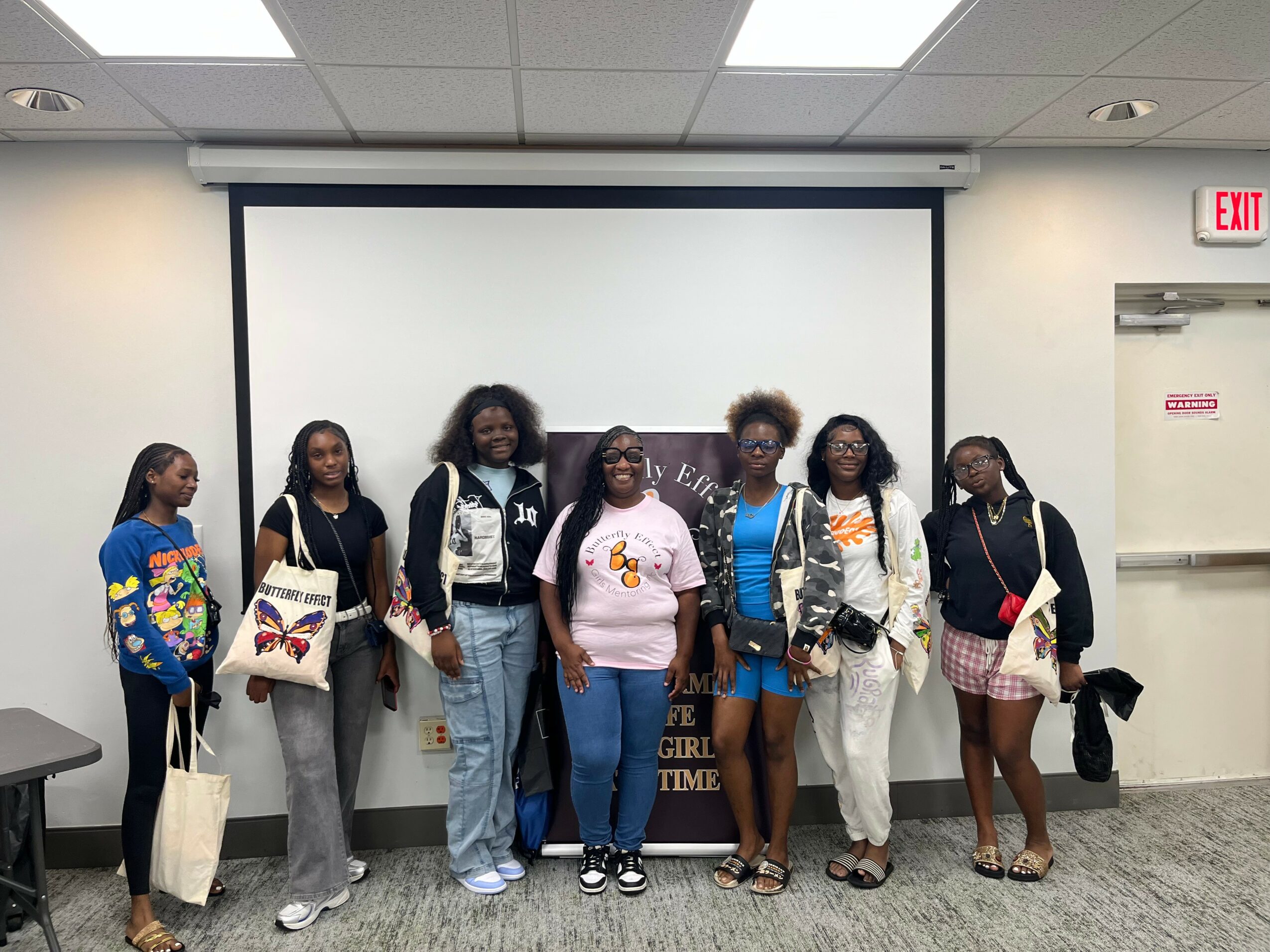 Six young women stand in front of a blank white screen in a classroom or conference room, holding bags and wearing casual clothing.