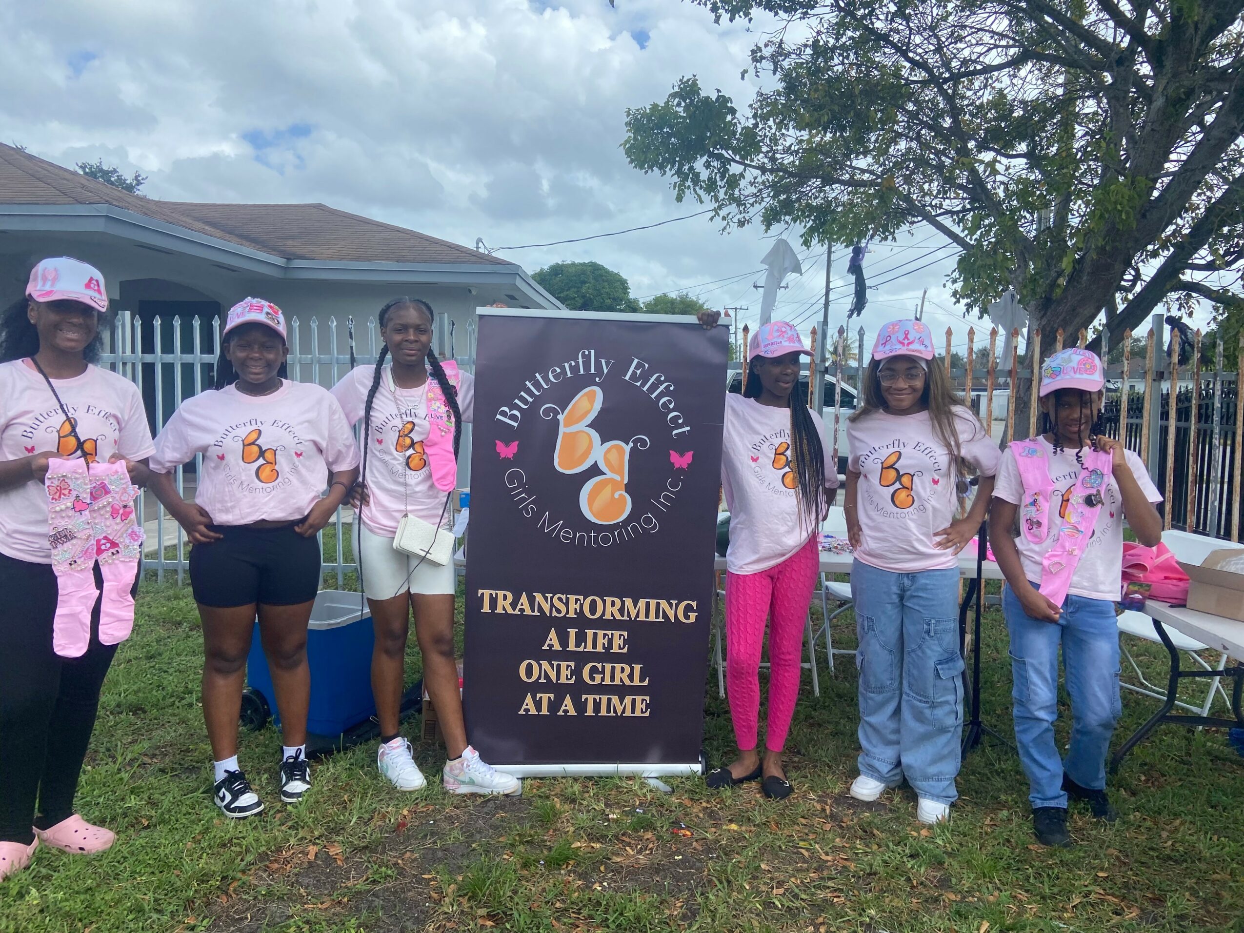 Group of seven girls wearing pink shirts and hats, standing outdoors with a butterfly-themed sign, trees, and a house in the background.