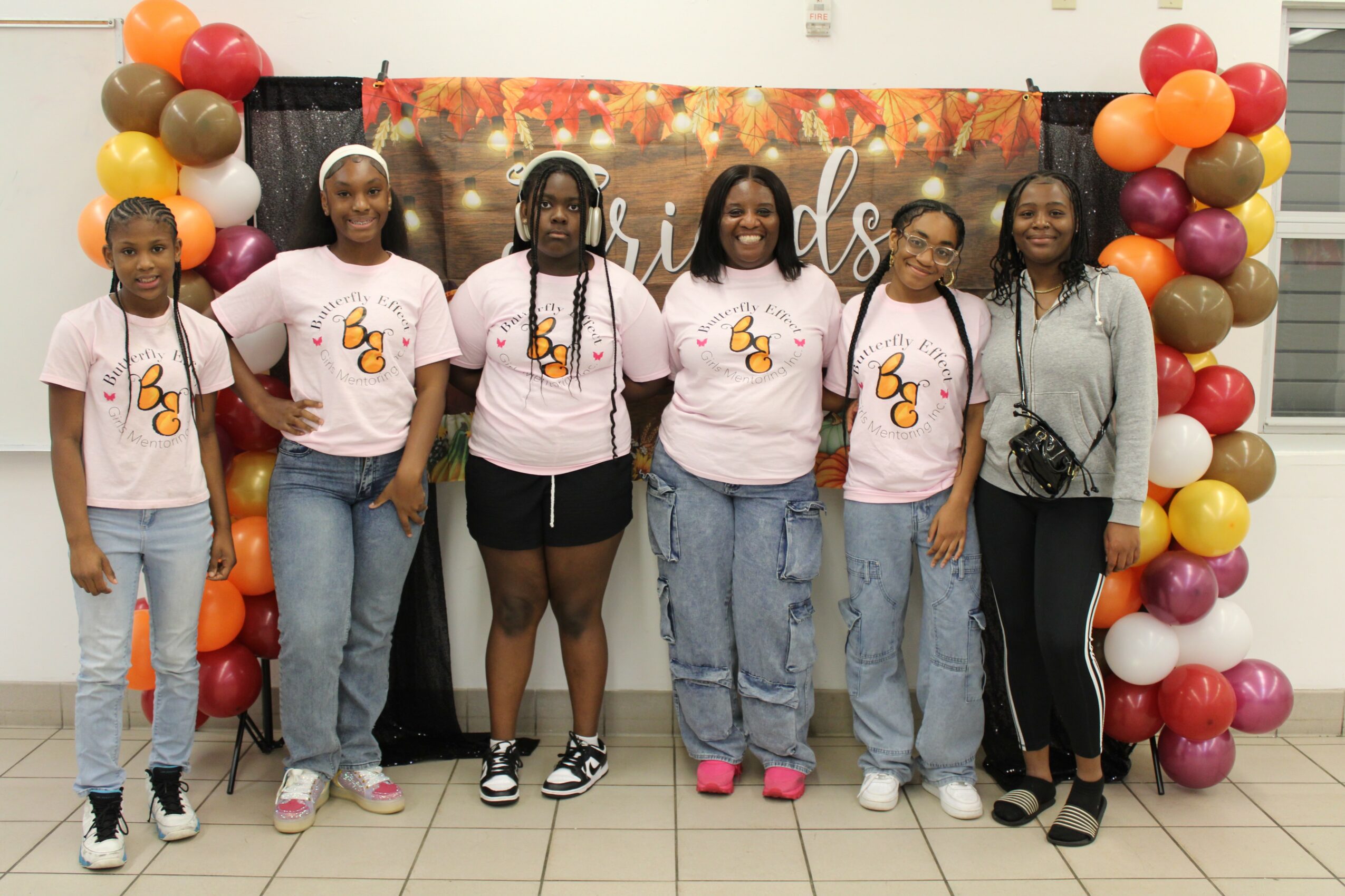 Six women stand in front of a colorful balloon arch and a decorated backdrop, smiling for a group photo.