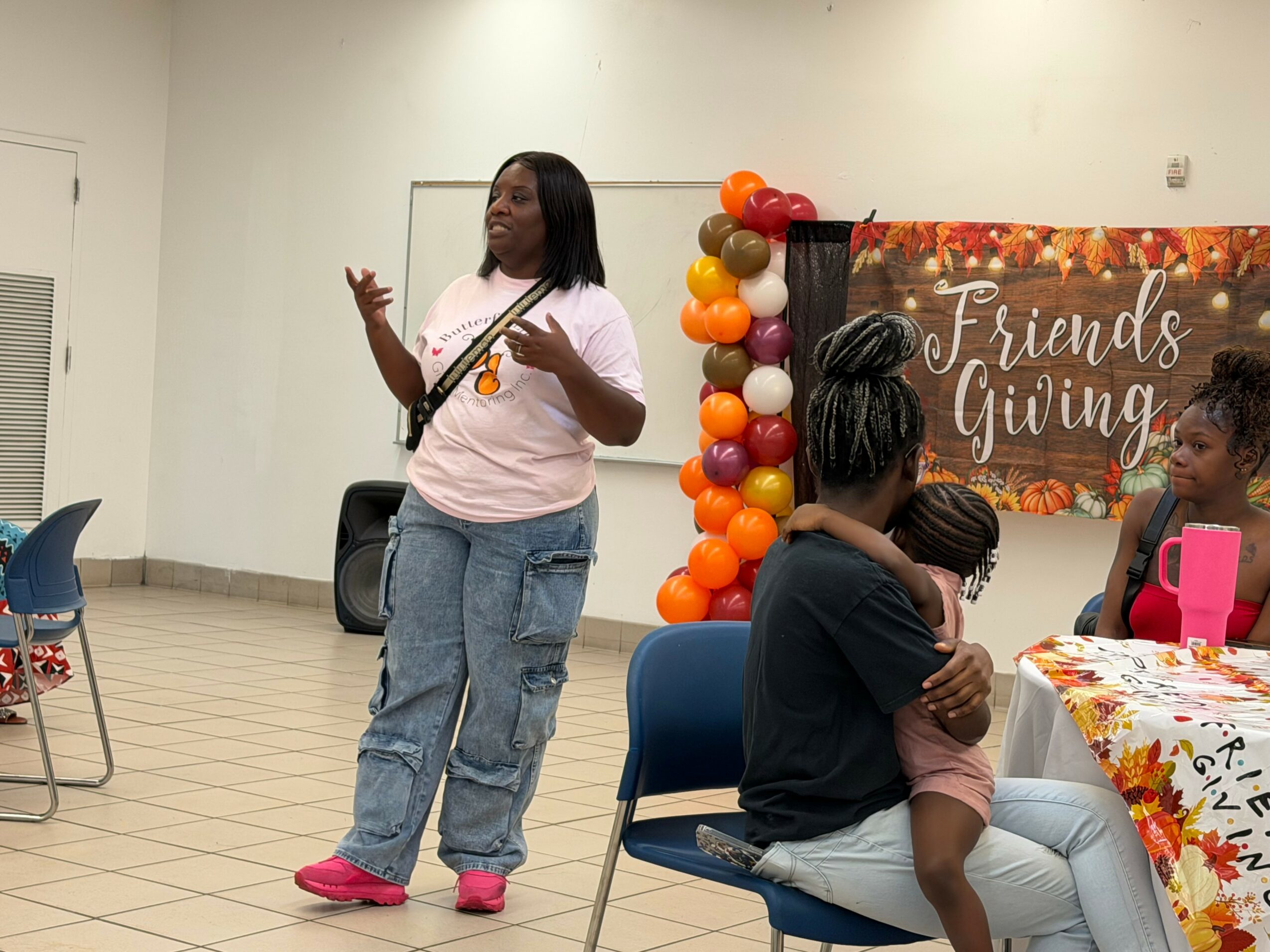 Woman speaking to a group at a Friendsgiving event, with a decorated balloon arch and table with a floral tablecloth.