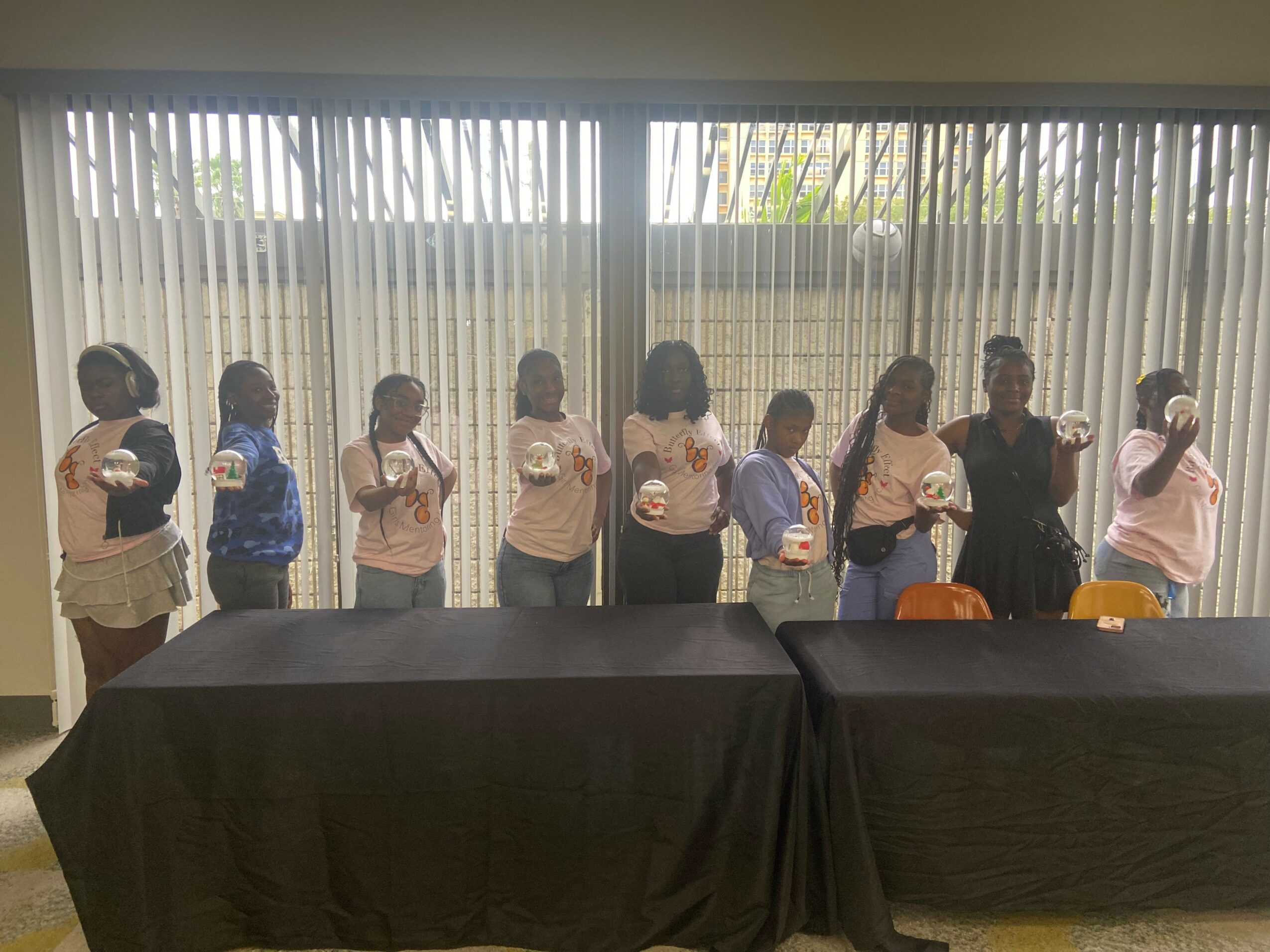 Group of children and two adults standing in line behind tables, facing forward, in front of vertical blinds.