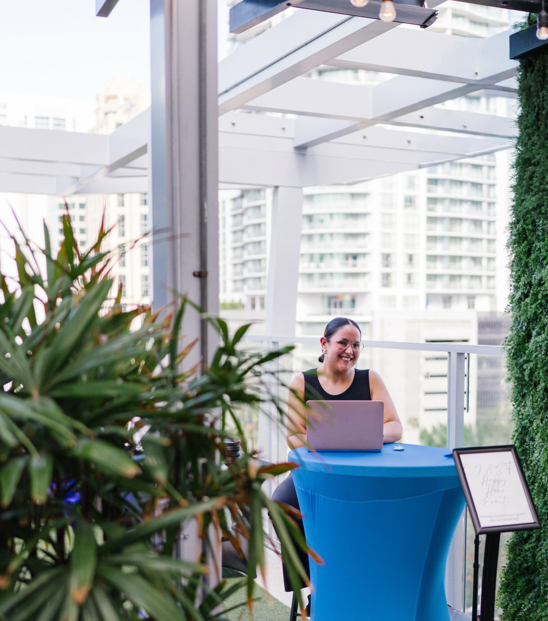 Woman sitting at a table with a laptop outdoors, with city buildings in the background.
