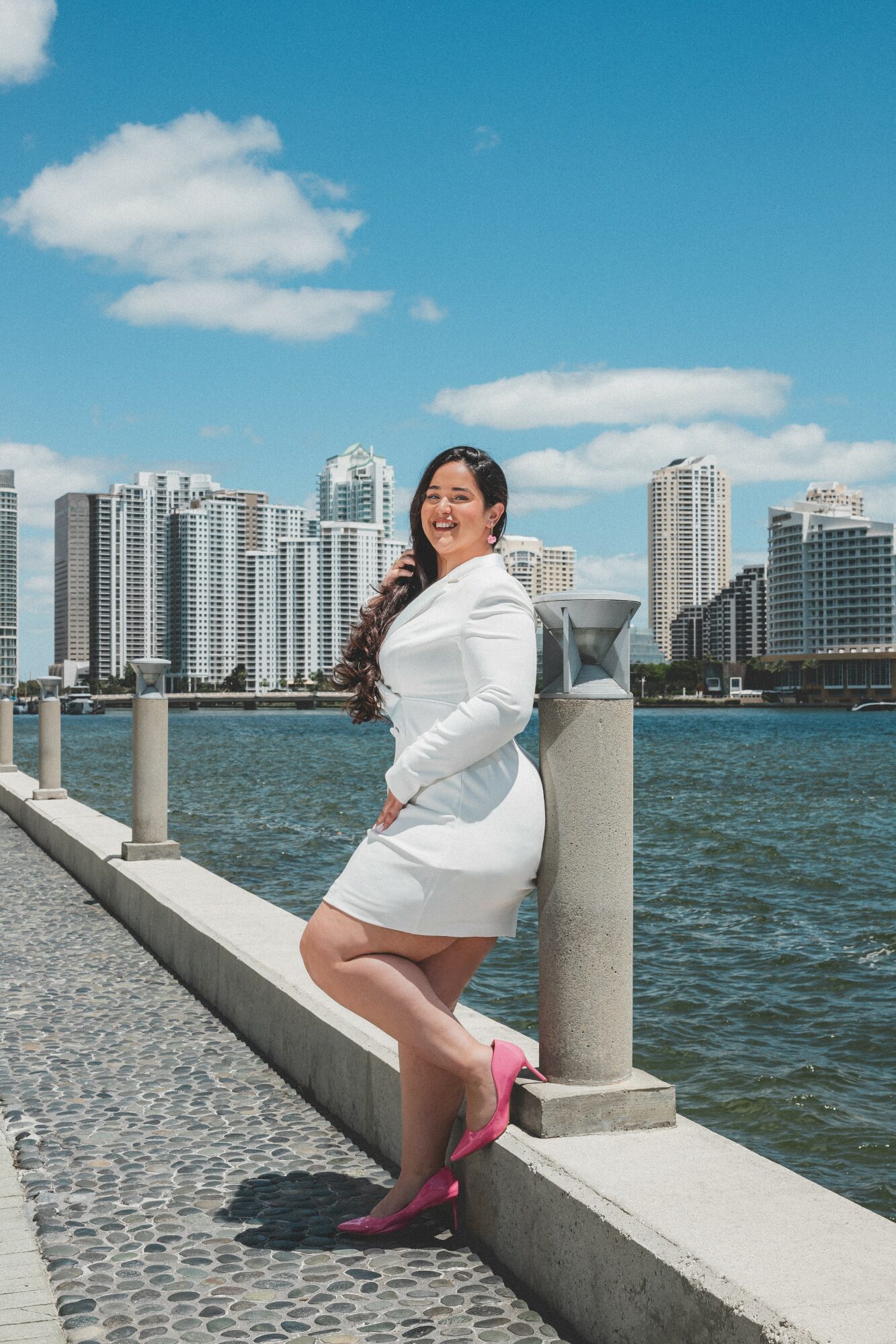 Woman in white coat and pink shoes standing by water with city skyline in background, smiling, sunny day.
