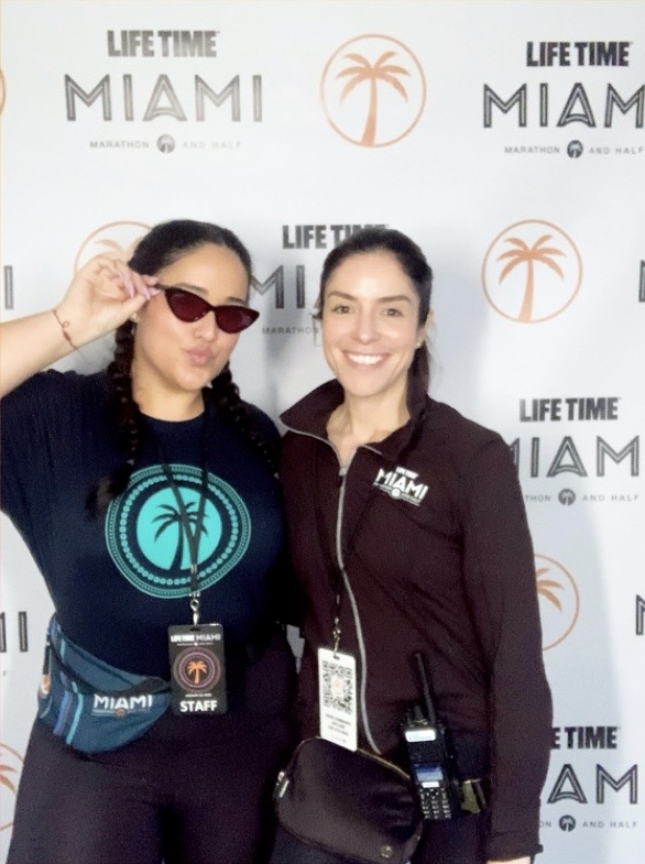 Two women smiling in front of a Miami-themed backdrop, one wearing sunglasses and a black jacket, the other in a black jacket with a badge.