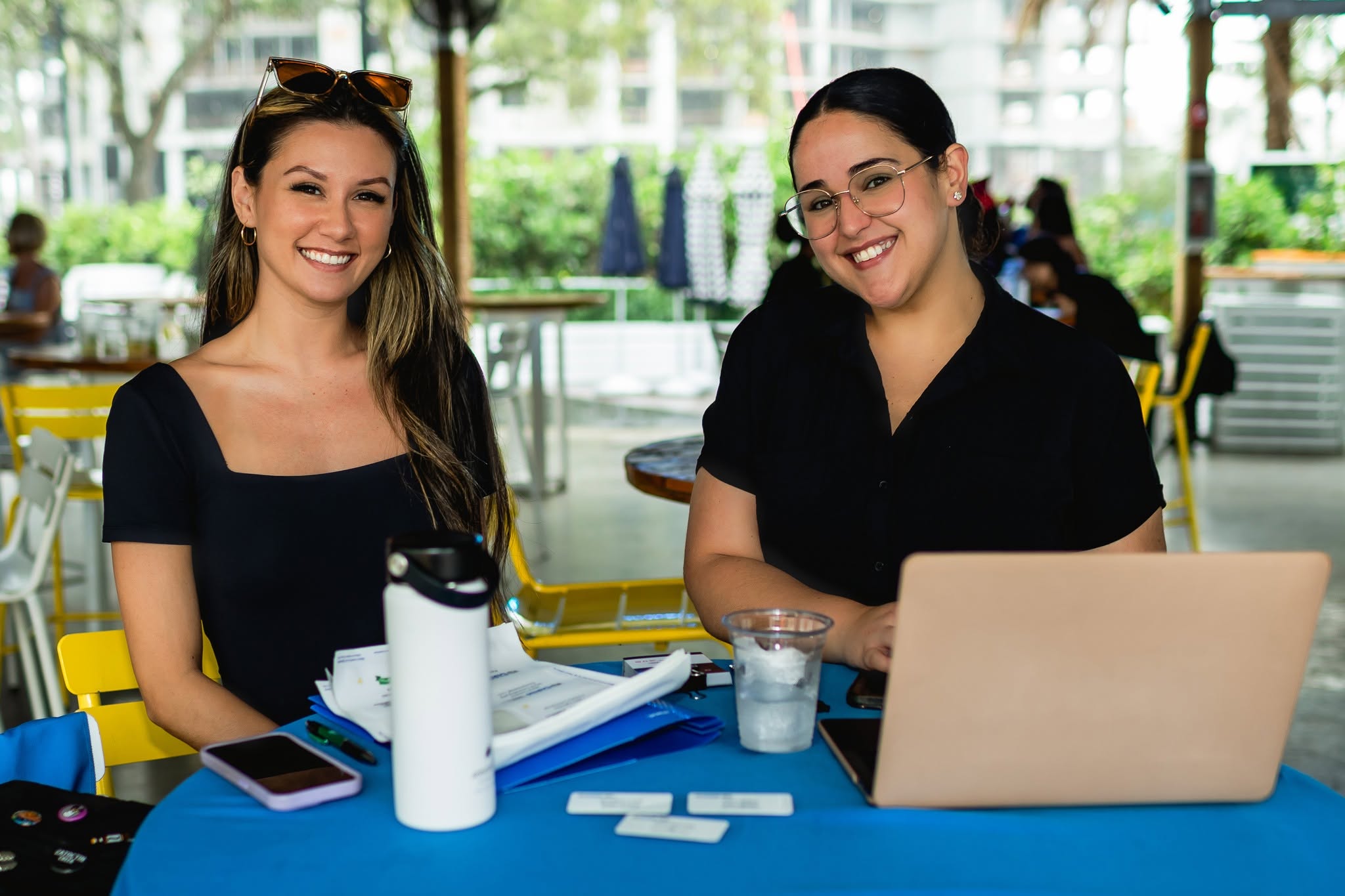 Two women sitting at a table outdoors, smiling, with a laptop, water bottle, and papers, in a bright setting.