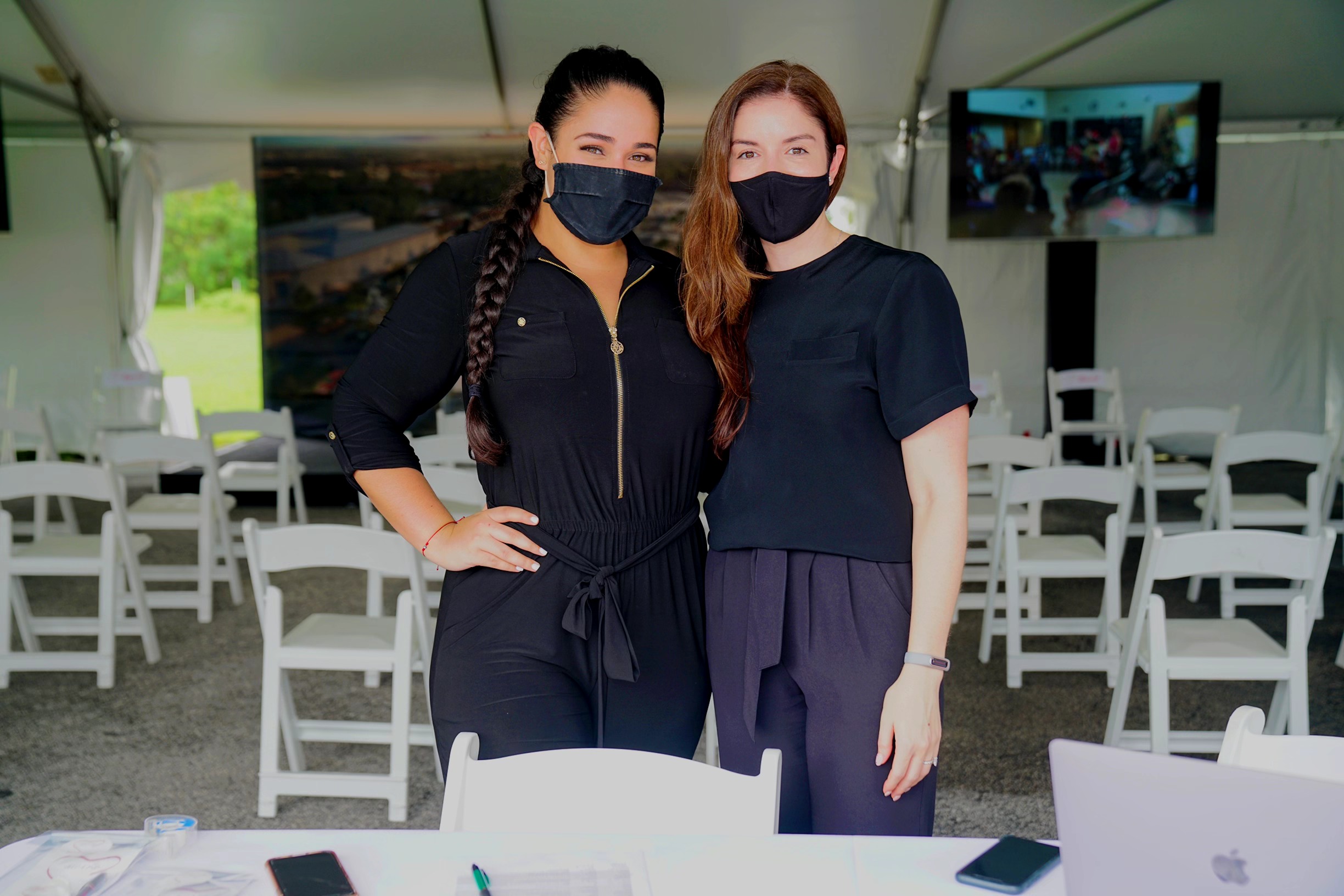 Two women wearing masks standing inside a tent with chairs and tables, large screens in the background.