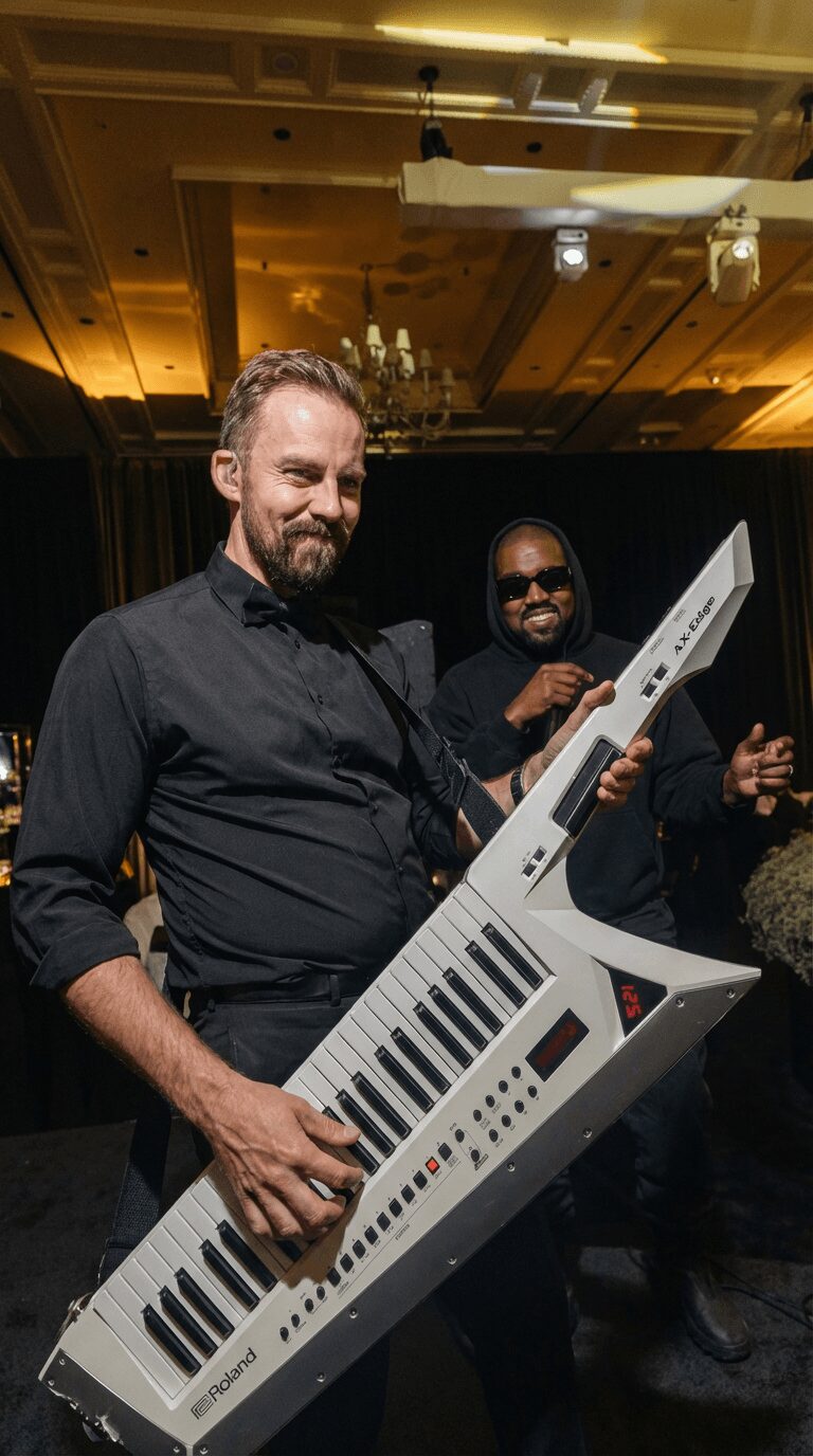 Two men at a party, one holding a large electronic keyboard, smiling, with a dark background and warm lighting.