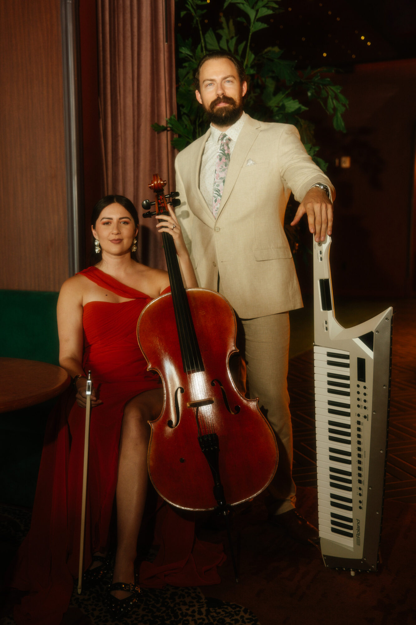A man in a beige suit and a woman in a red dress pose with musical instruments indoors, with a dark background and green plants.