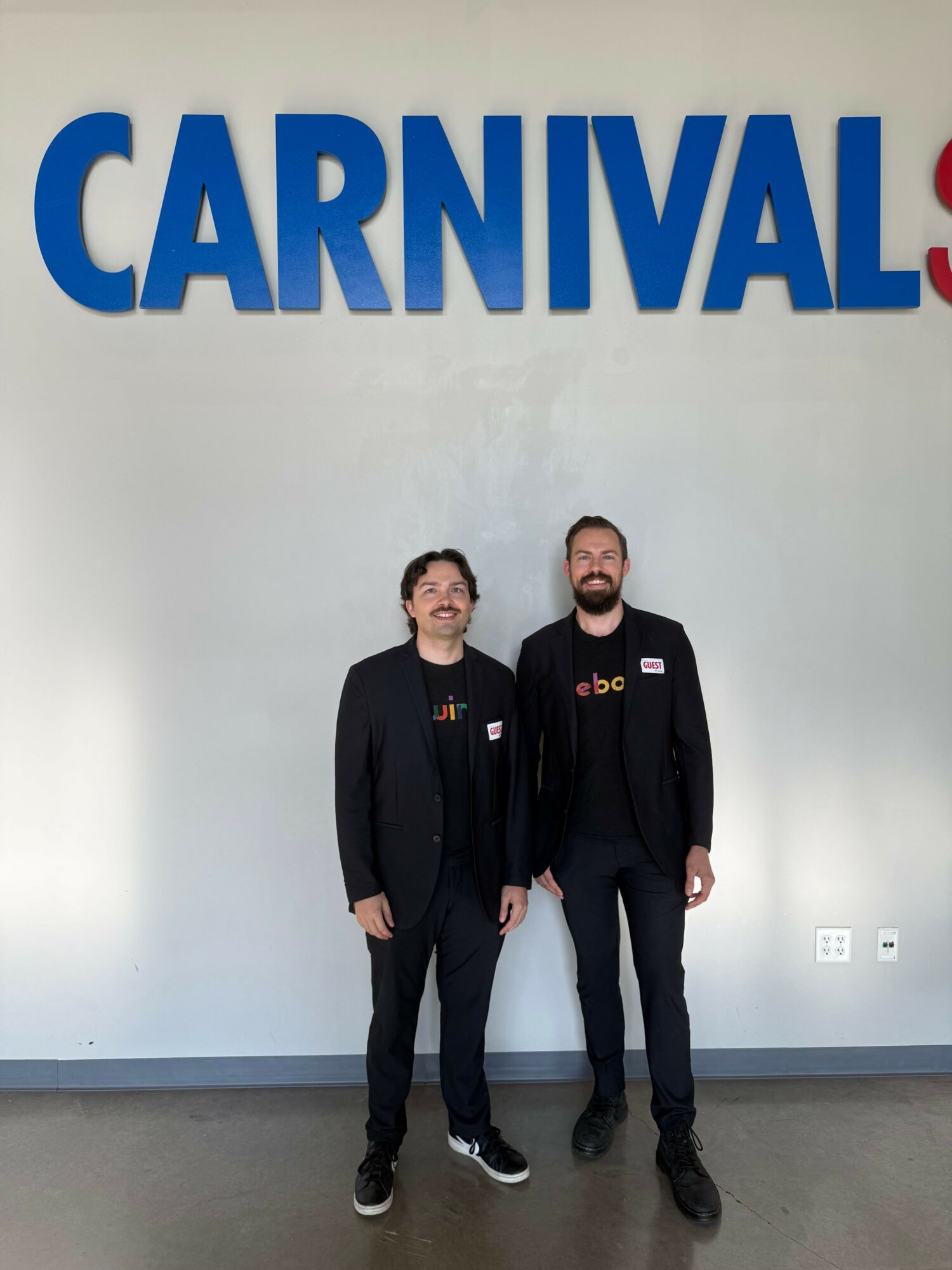 Two men in black suits standing in front of a wall with large blue and red text reading 'CARNIVAL'.