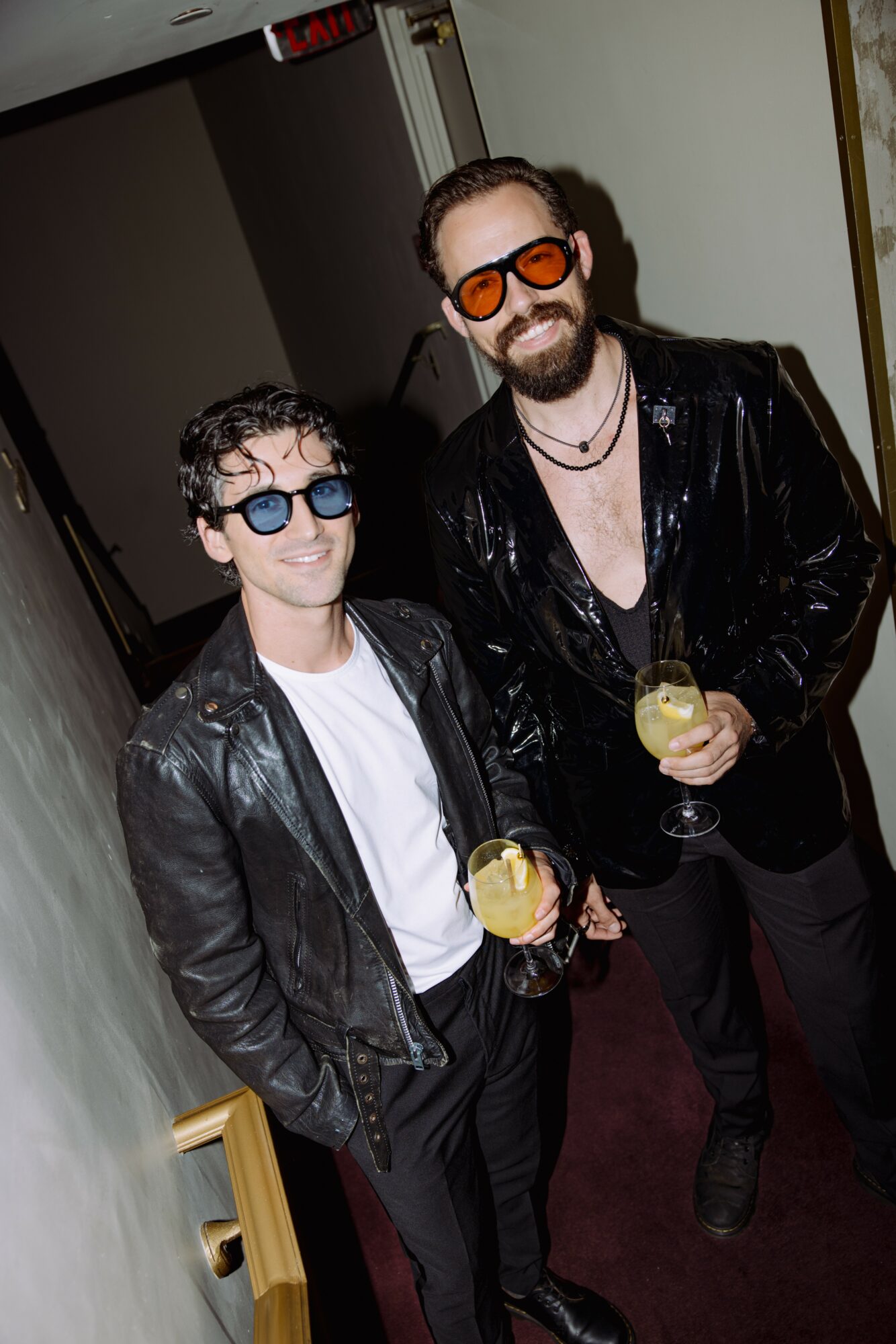 Two men wearing sunglasses and leather jackets holding drinks, standing indoors with a dark background.