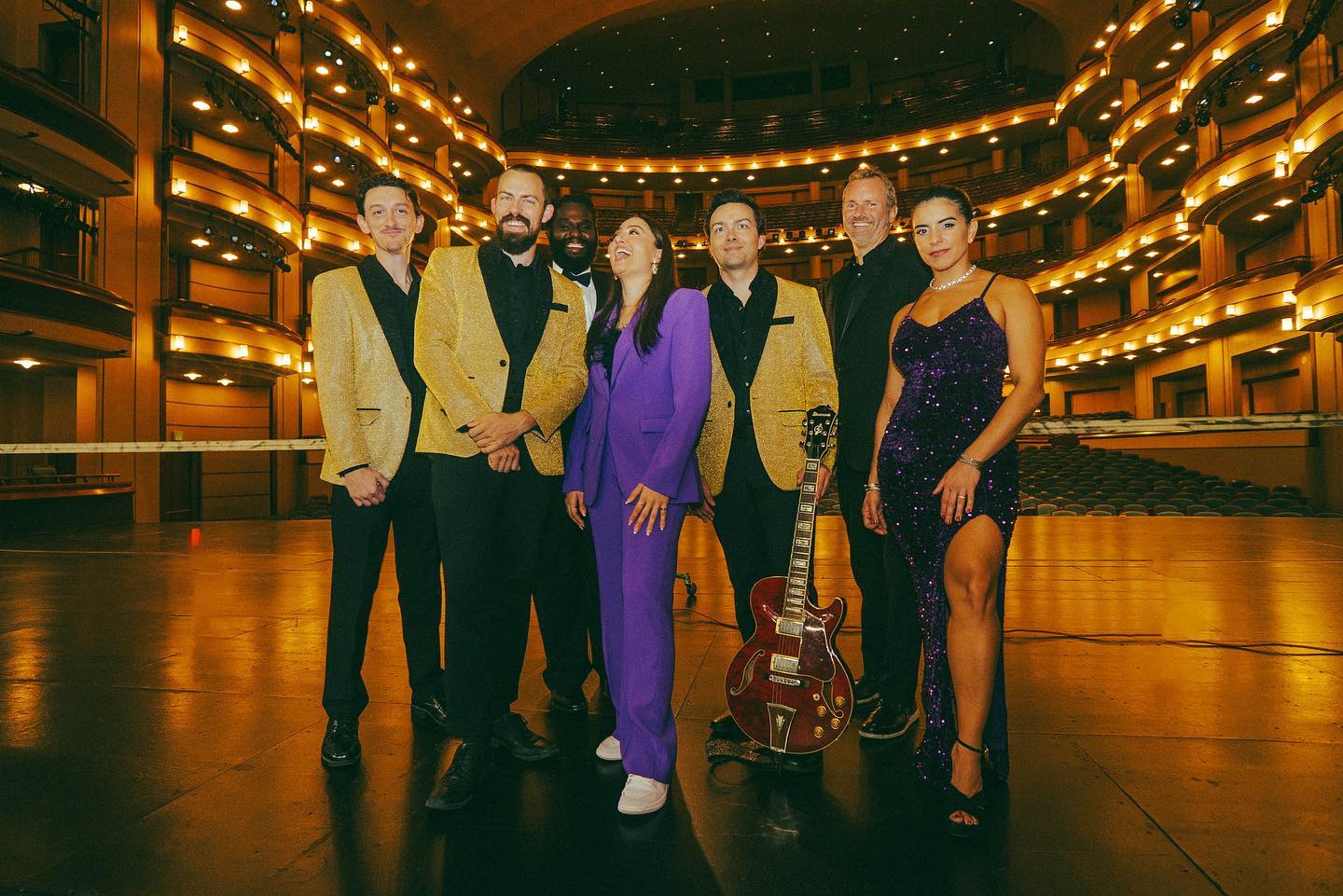 Group of five people standing on stage in a theater with balconies and warm lighting, dressed in formal and performance attire.
