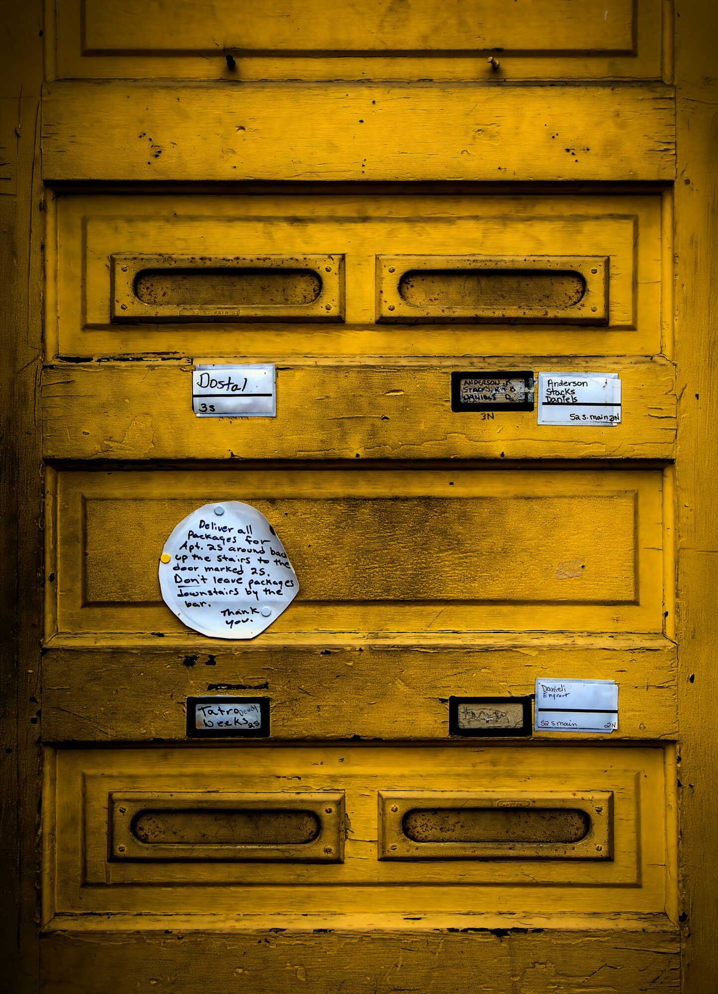 Yellow mailbox with various labels and a round note attached, showing signs of wear and weathering.