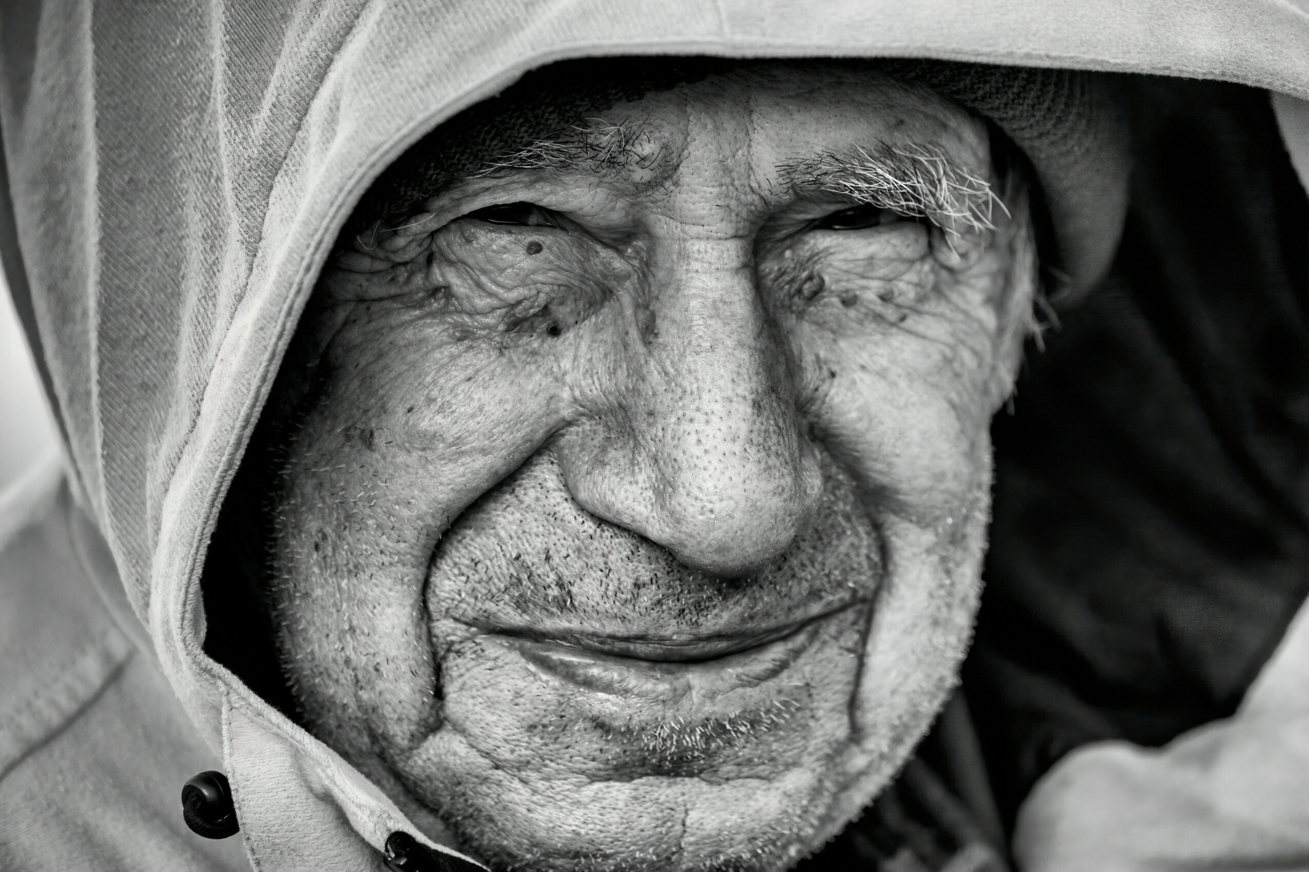 Close-up of an elderly person's face with a hood, smiling gently, showing wrinkles and textured skin.