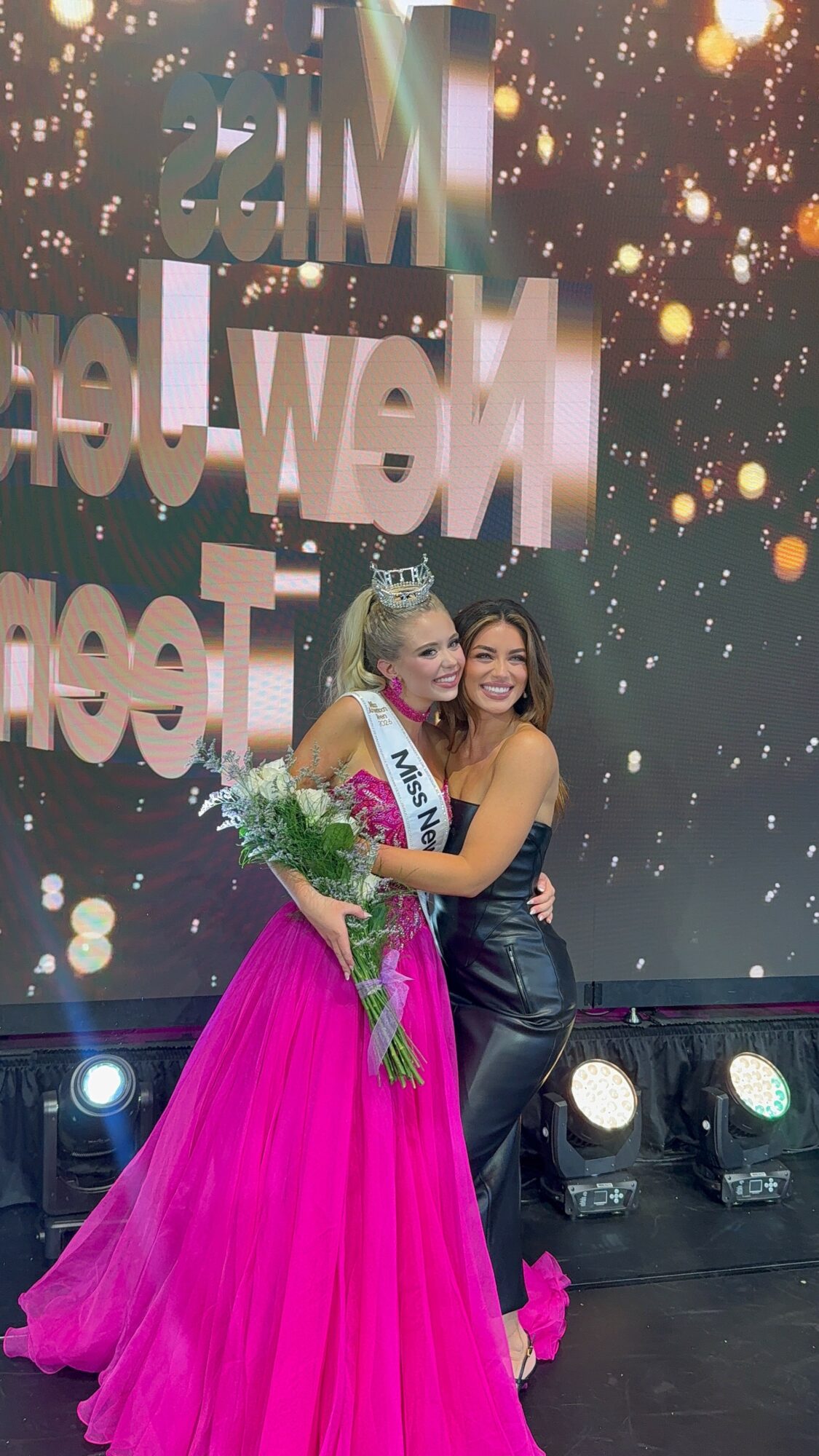 Two women on stage, one in a pink gown and crown, holding a bouquet, smiling together.