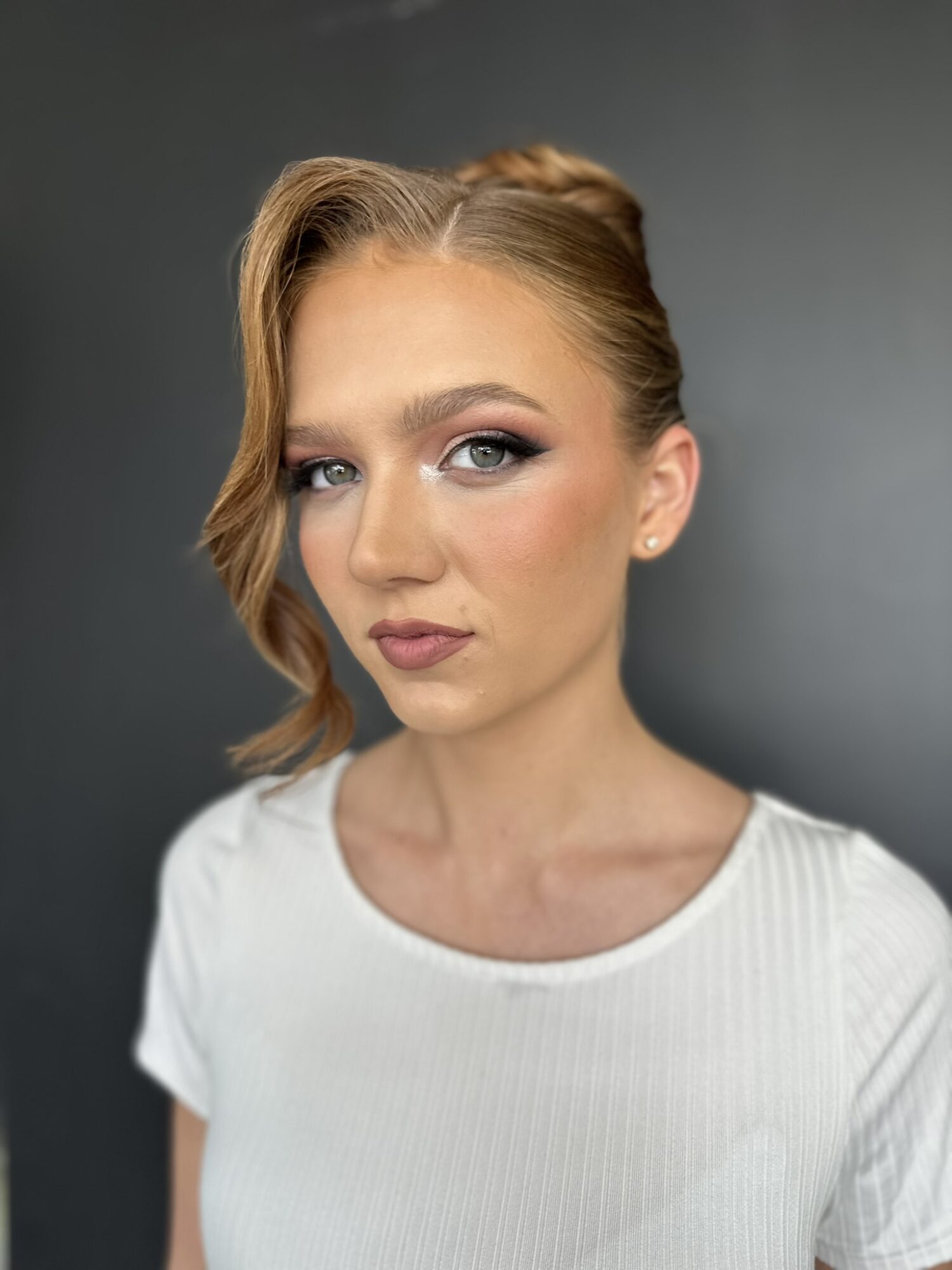 Portrait of a young woman with styled hair, makeup, and a white top, looking at the camera against a dark background.