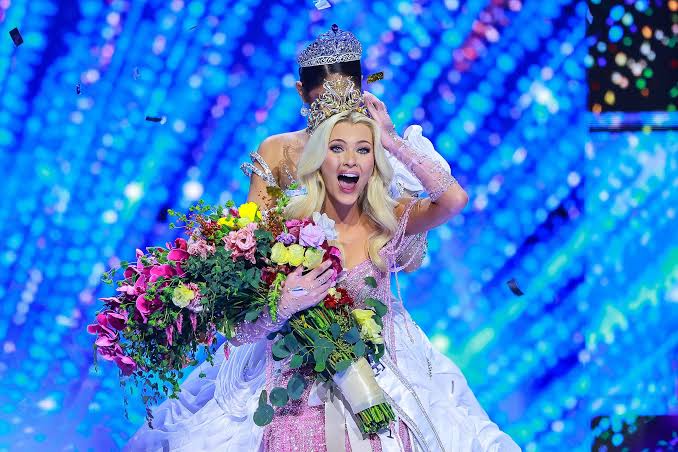 A woman in a white dress holds a large bouquet of flowers, smiling with a person behind her, against a blue background.
