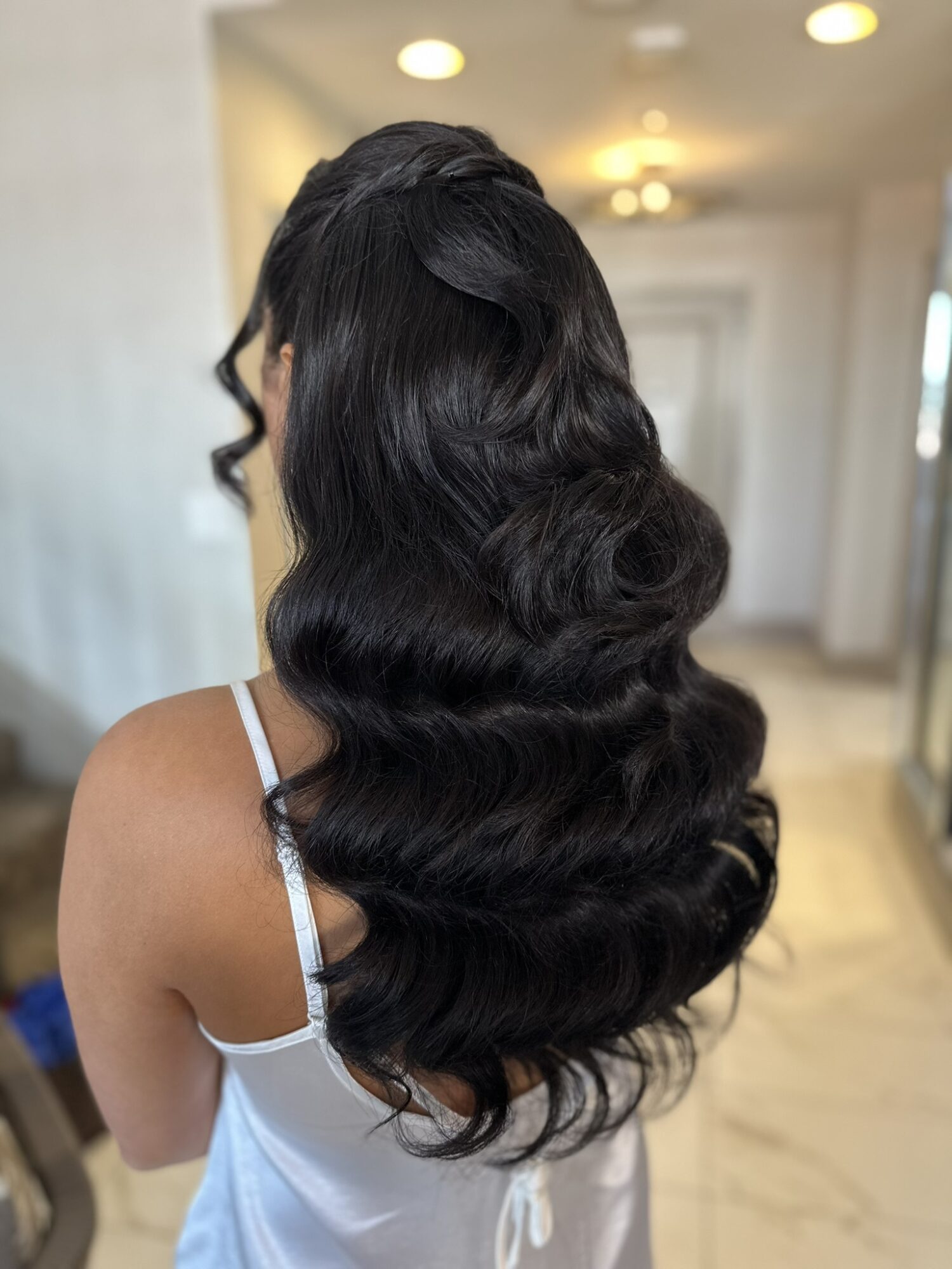 Back view of a woman with long, wavy black hair in a well-lit room, wearing a white top with thin straps.