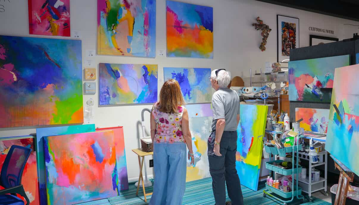 Two people in an art studio surrounded by colorful abstract paintings and art supplies.