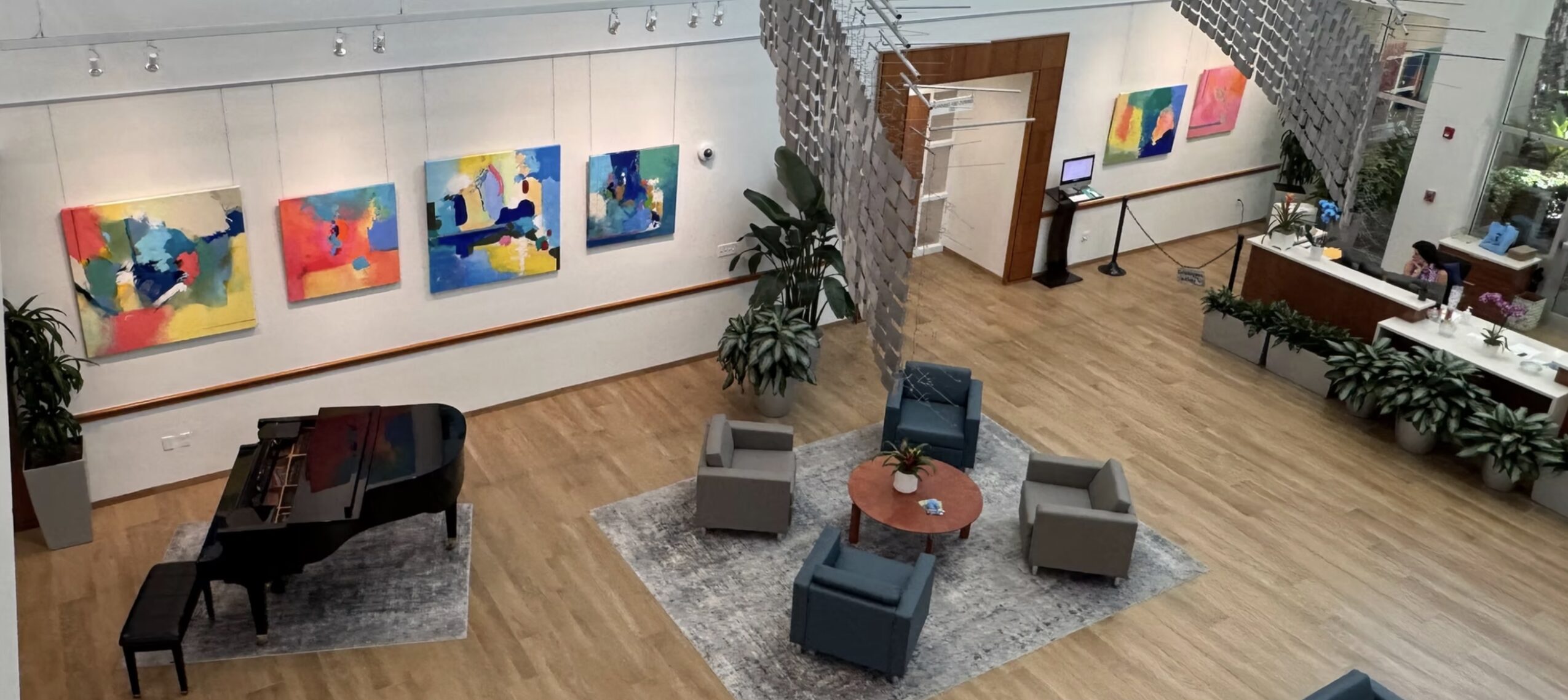 Art gallery with colorful paintings on white walls, a piano, and a seating area with chairs and a table.