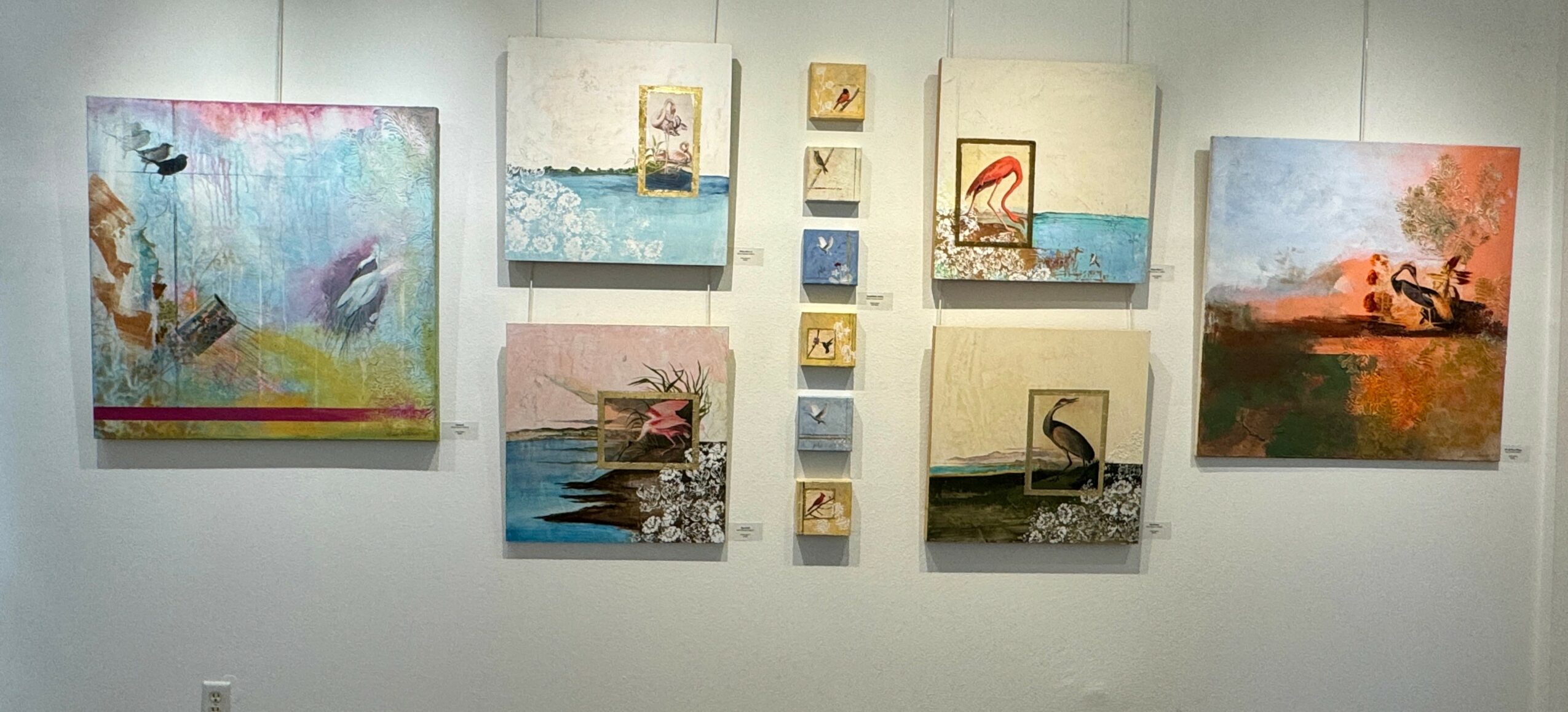 Gallery wall with abstract and landscape paintings, some with water themes, arranged with small square artworks in the center.