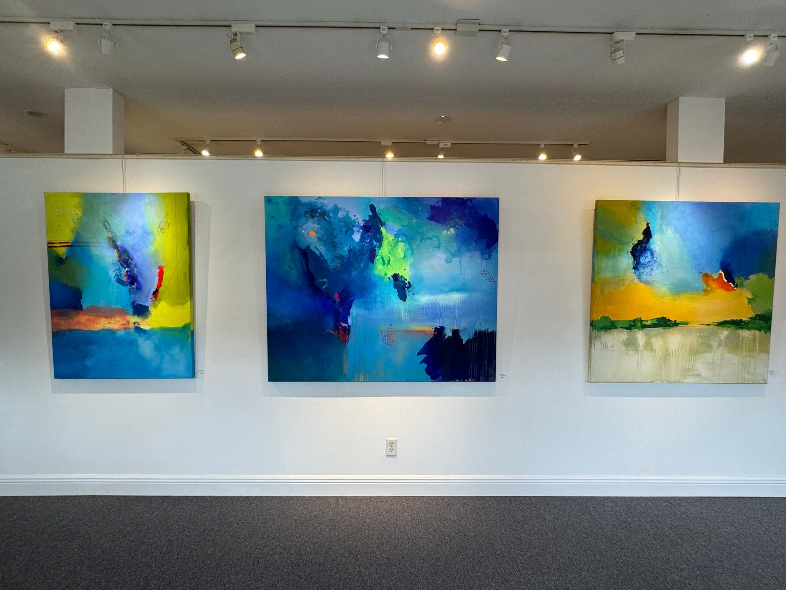 Three abstract paintings with vibrant colors hang on a white gallery wall, illuminated by ceiling lights.