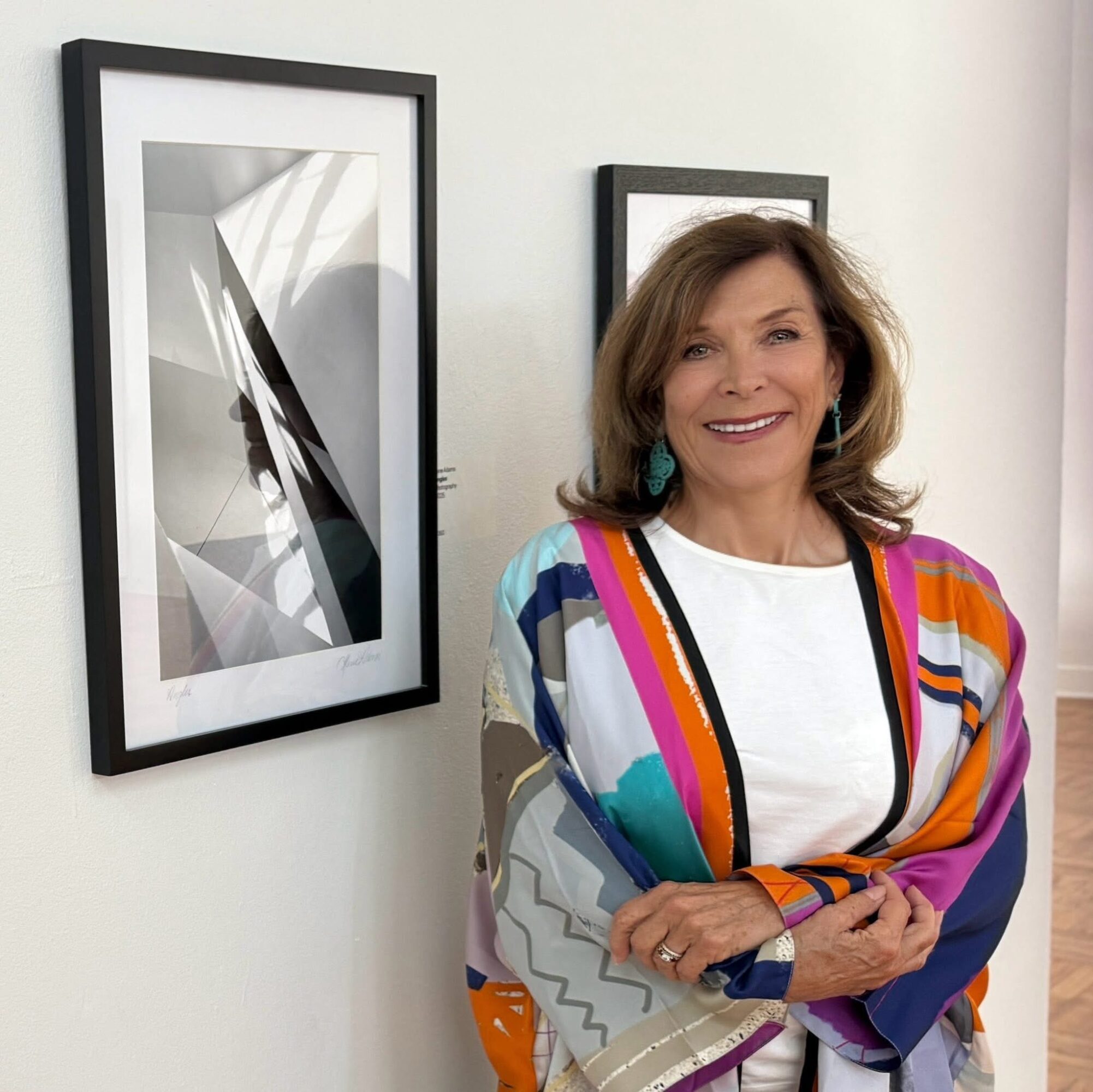 Woman with shoulder-length hair smiling, wearing a colorful striped shawl, standing next to black-framed abstract artwork on white wall.