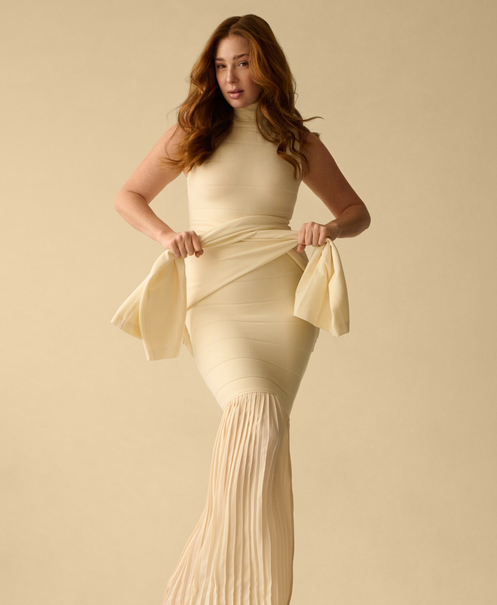 Woman with long wavy hair wearing a cream-colored dress holding a matching jacket, standing against a plain background.