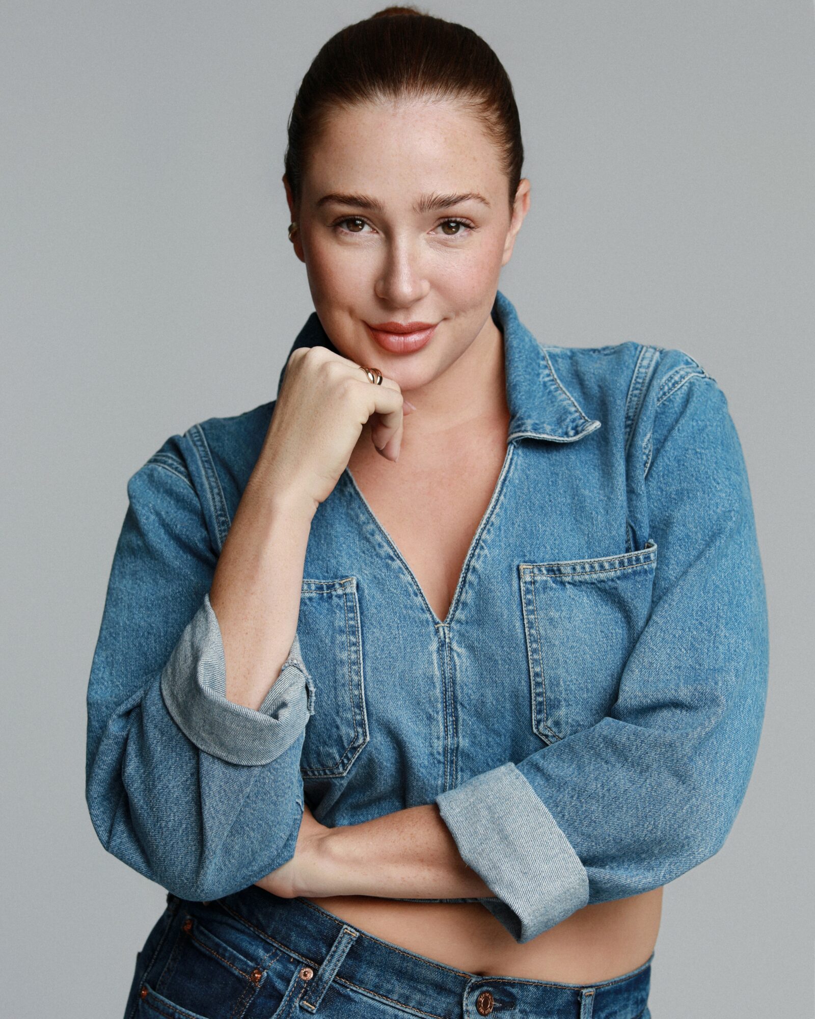 Young woman with red hair in a denim jacket, resting her chin on her hand, smiling slightly, against a plain gray background.